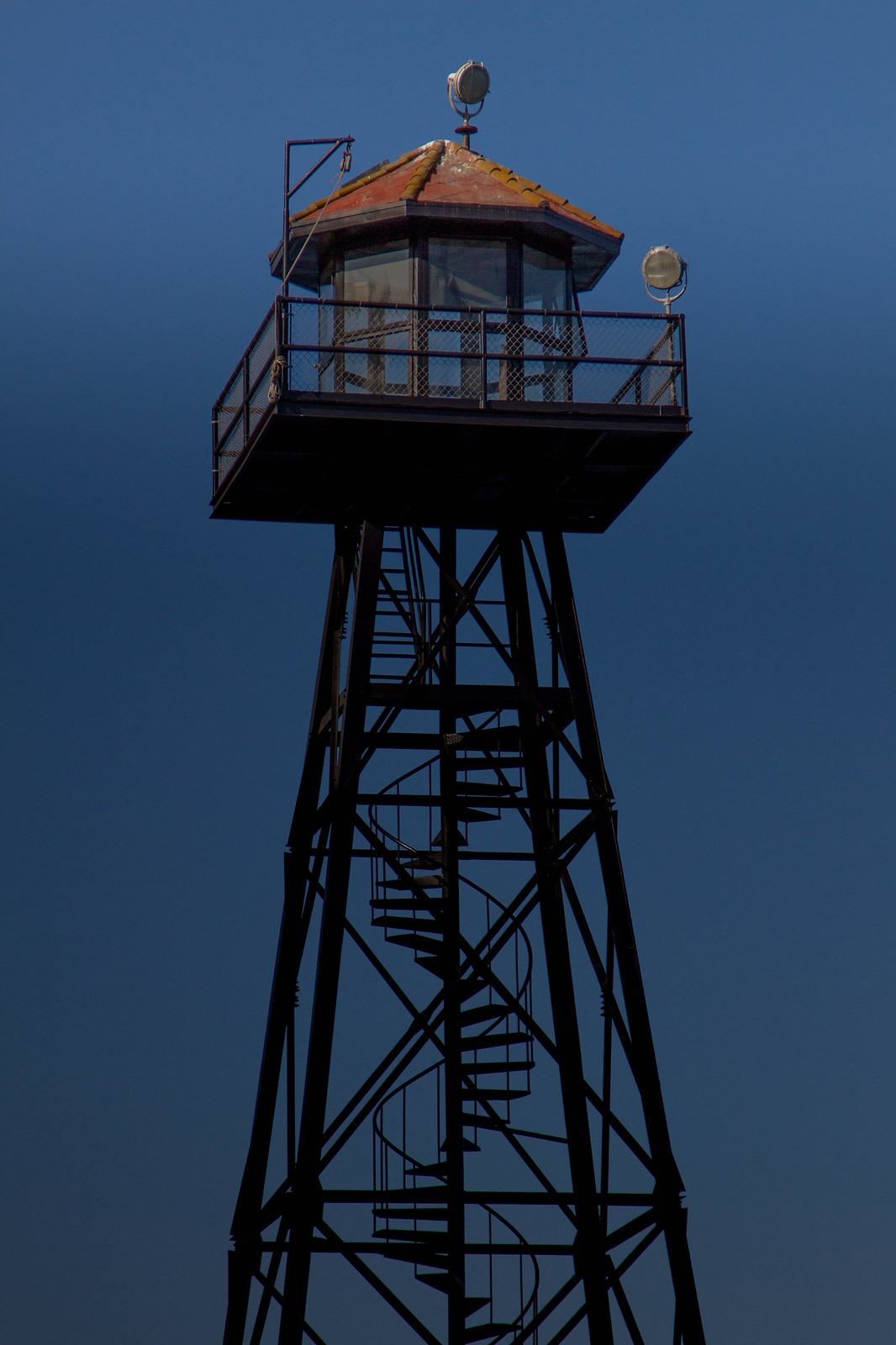 Silhouetted Alcatraz guard tower with a spiral staircase and searchlights against a deep blue sky.