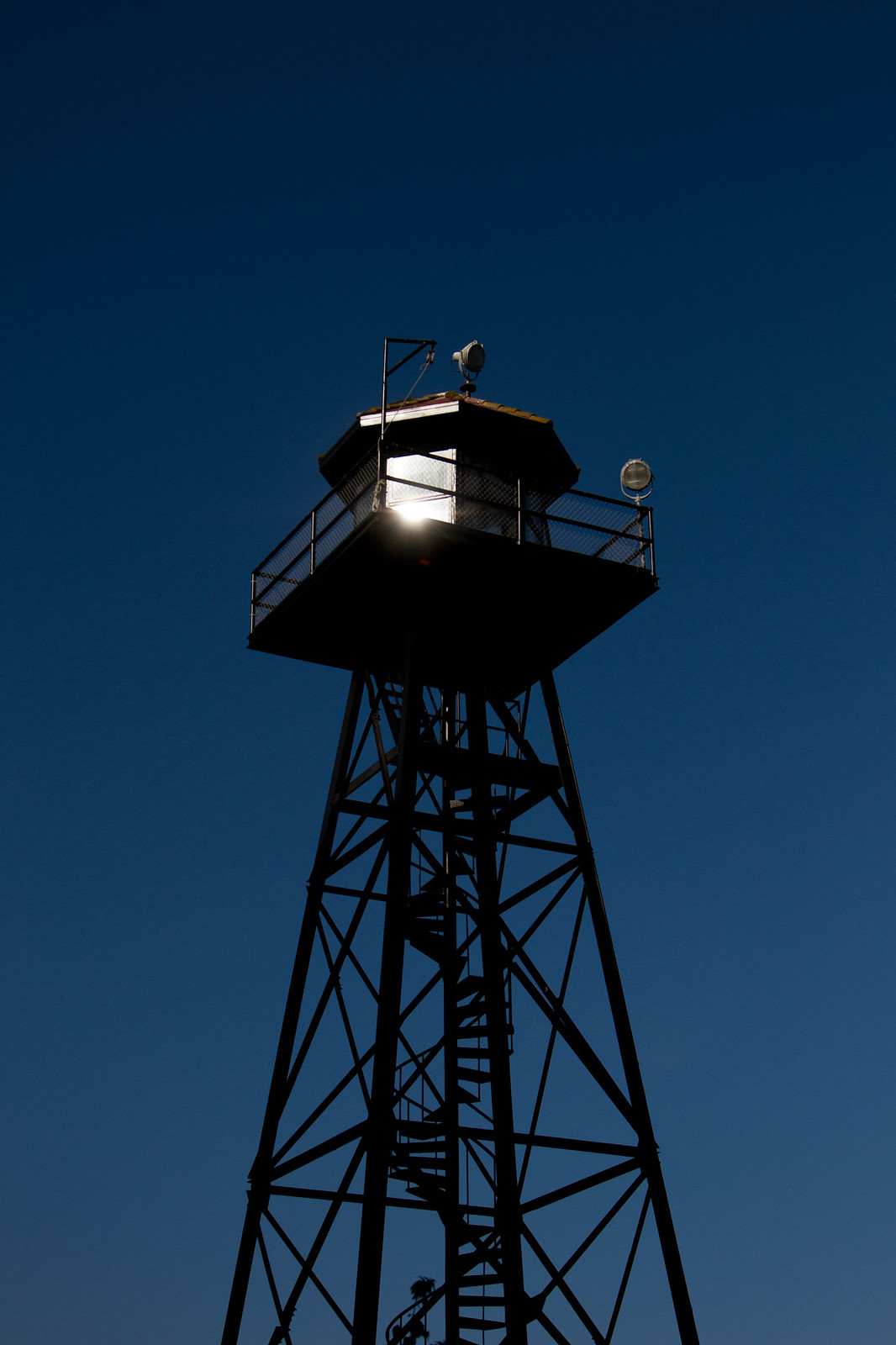 Silhouetted Alcatraz guard tower with searchlights against a deep blue sky, sun flaring through its windows.