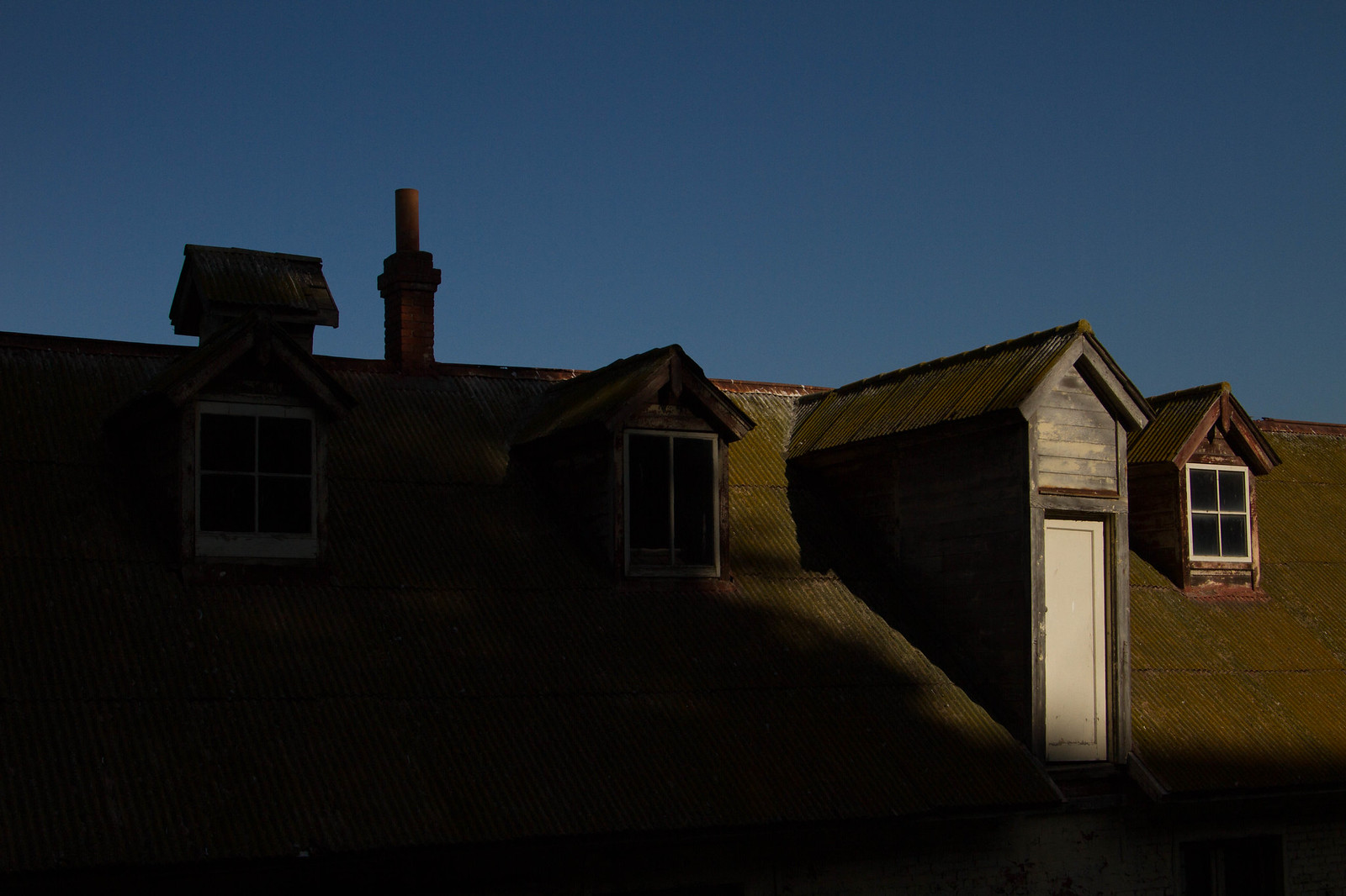 Weathered dormer windows and a brick chimney rise from a moss-covered corrugated metal roof under a deep blue sky.