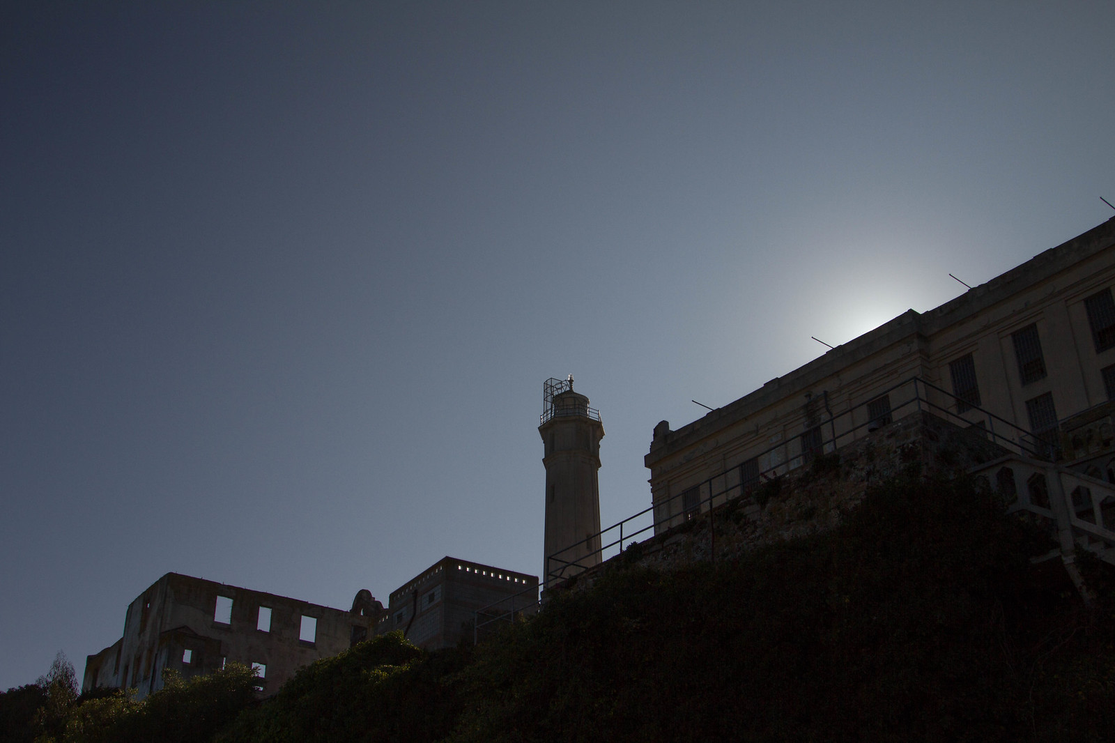 The Alcatraz lighthouse and cellhouse silhouetted against a dusk sky, viewed upward from below the island's hillside.