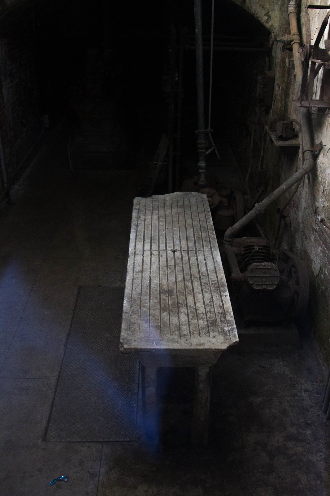 Grooved concrete morgue slab beside rusted machinery in a dark, brick-walled basement room at Alcatraz.