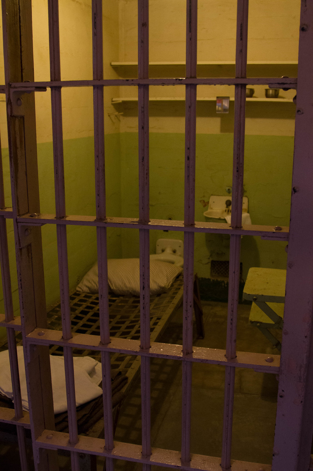 View through worn purple bars into an Alcatraz cell with a folded bunk, sink, and green-and-white walls.