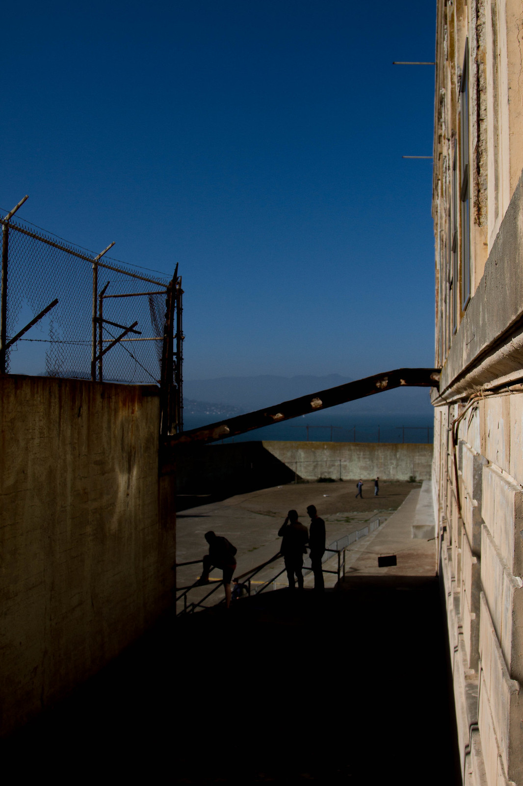 Silhouetted visitors in Alcatraz's walled recreation yard, framed by barbed-wire fencing and the bay beyond.