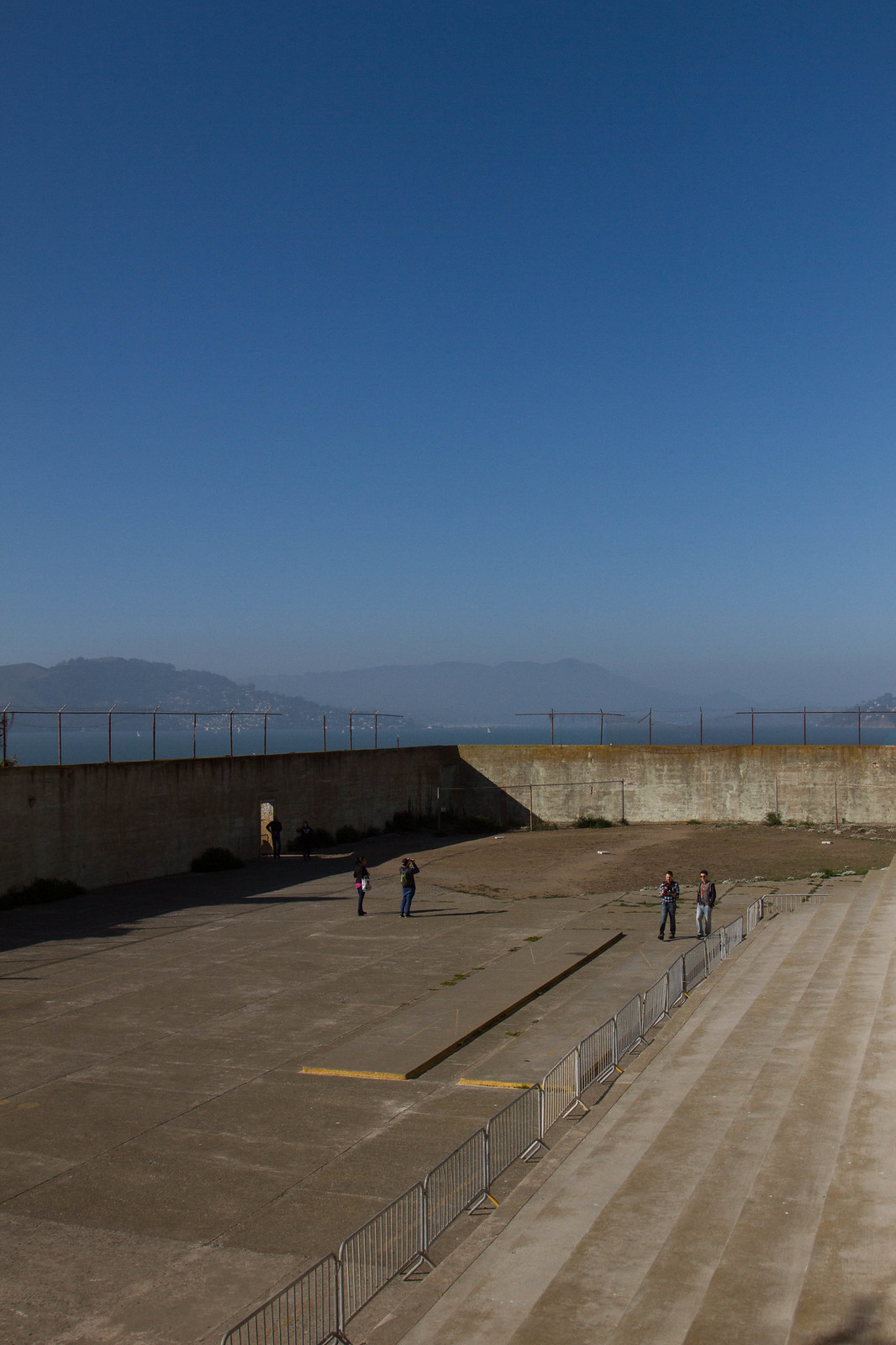 Visitors stand in Alcatraz's empty concrete recreation yard, its high walls topped with fencing above San Francisco Bay.