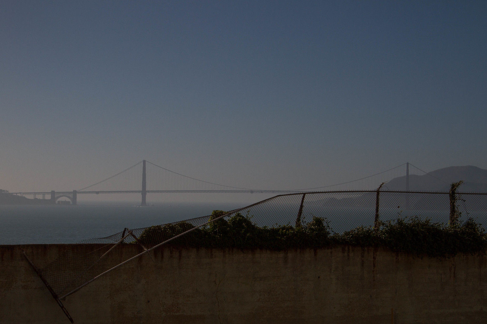 The hazy Golden Gate Bridge seen across the bay over a broken chain-link fence and crumbling concrete wall.