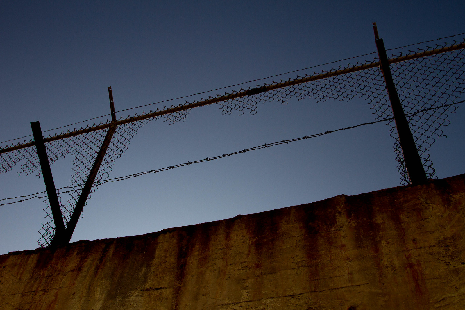 Barbed-wire-topped chain-link fence silhouetted atop a weathered concrete prison wall against a deep blue dusk sky.