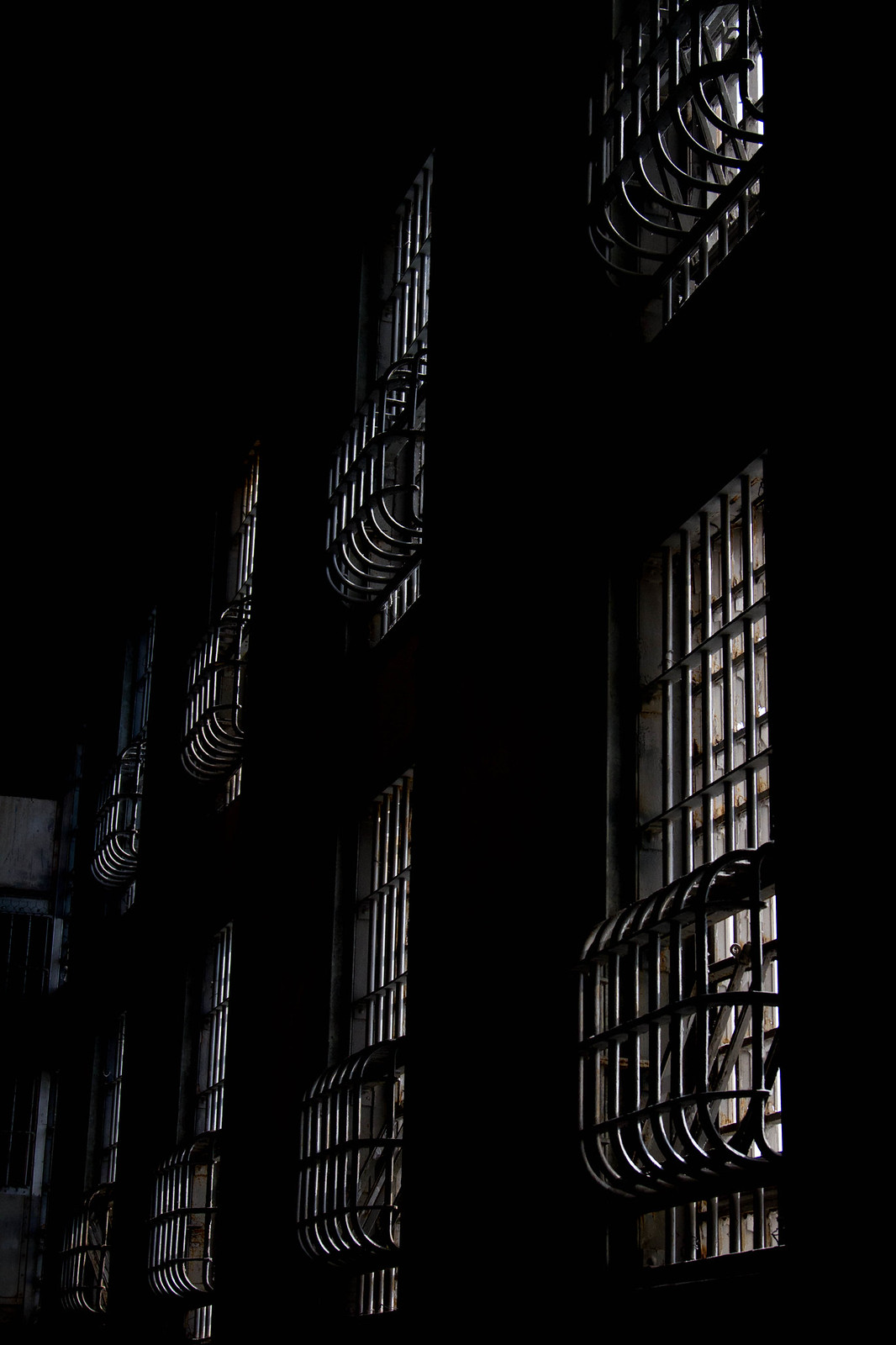 Barred windows with curved metal cages catch dim light against the dark interior wall of Alcatraz prison.
