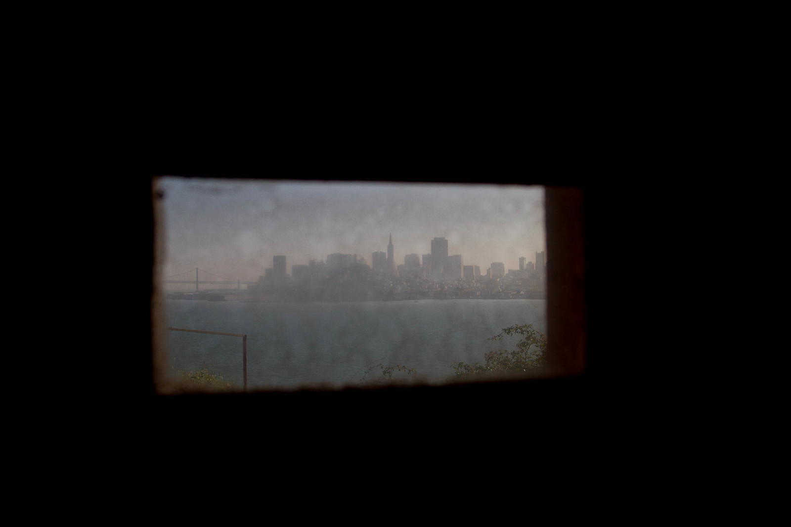 The San Francisco skyline and Bay Bridge in haze, framed through a dark grimy Alcatraz window.