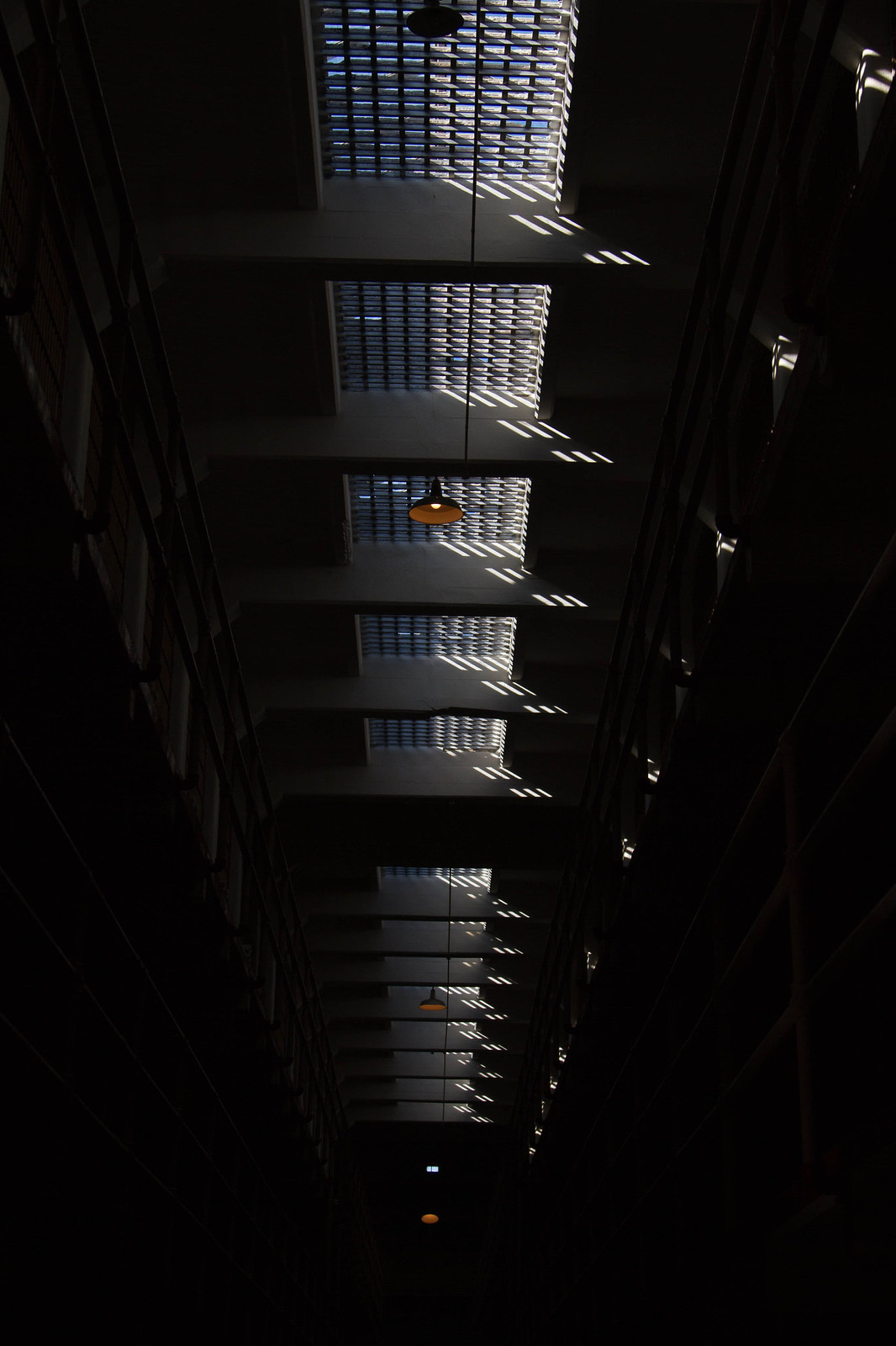 Daylight streams through a row of barred skylights above a dark cellblock, past hanging pendant lamps.
