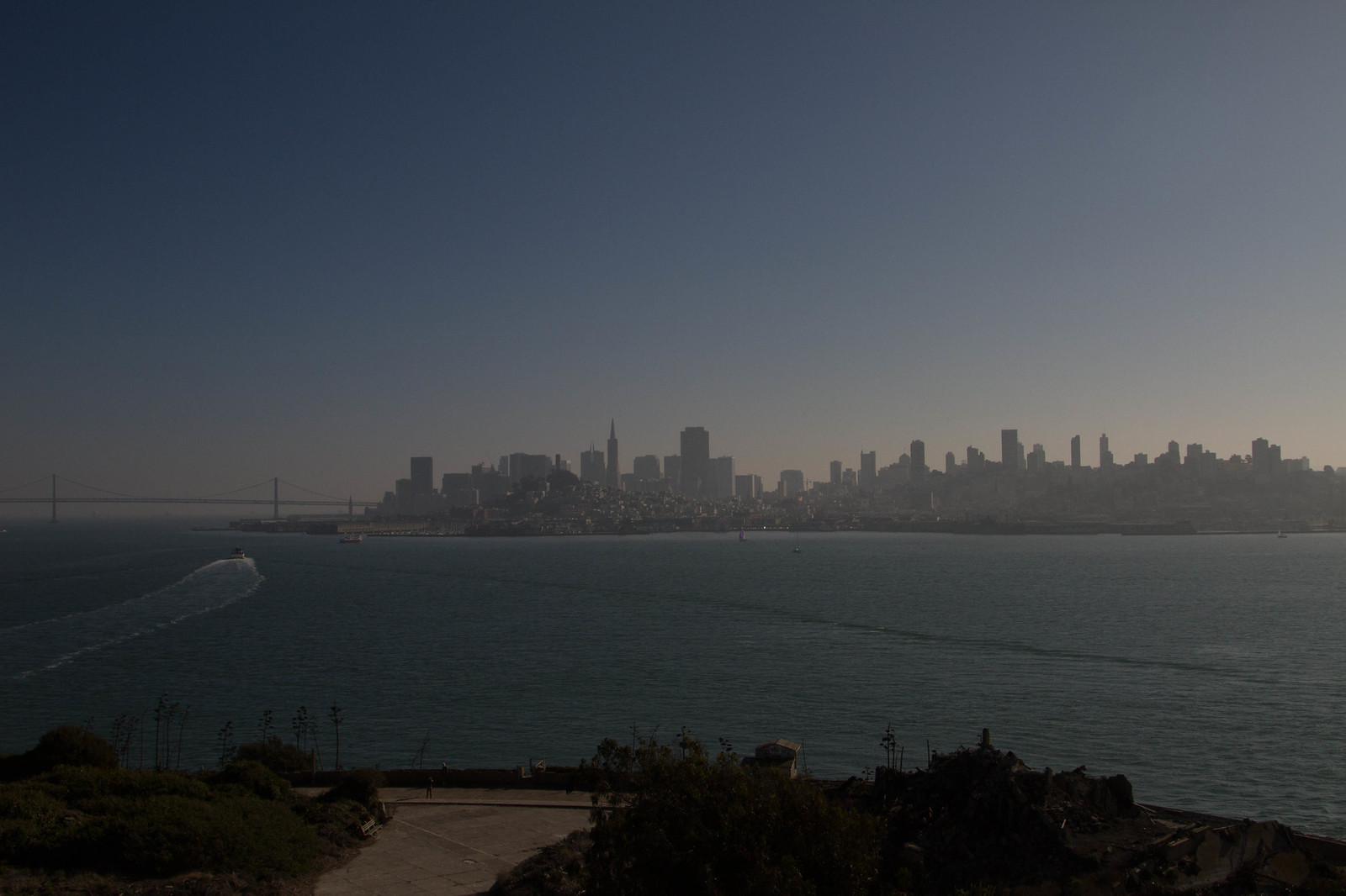The hazy San Francisco skyline and Bay Bridge seen across the water from Alcatraz Island, a boat trailing a wake.