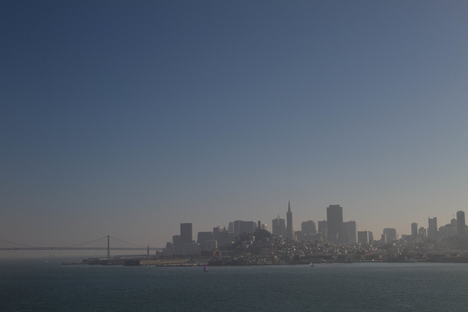 Hazy San Francisco skyline seen across the bay, with the Bay Bridge and Transamerica Pyramid under a clear sky.