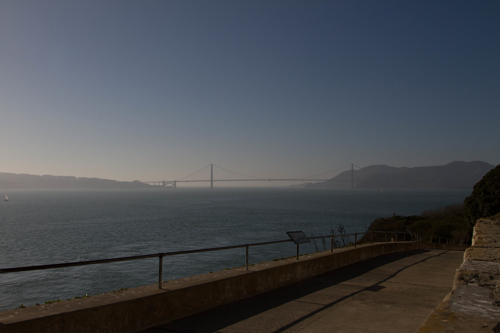 The Golden Gate Bridge in hazy light across the bay, seen from a paved walkway with a railing and an interpretive sign on Alcatraz.