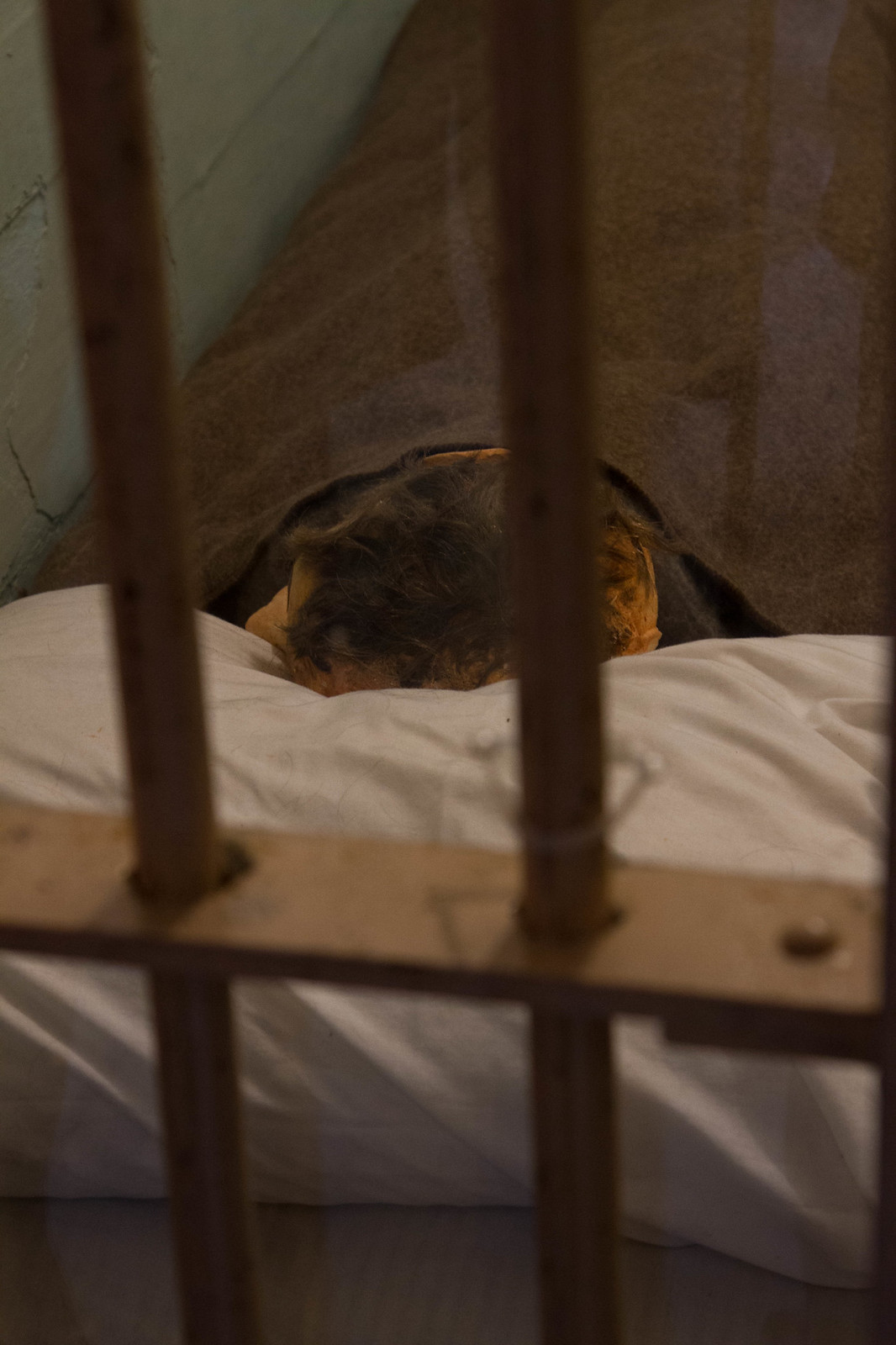 A dummy head with real hair tucked under a blanket in an Alcatraz cell bed, seen through the bars.