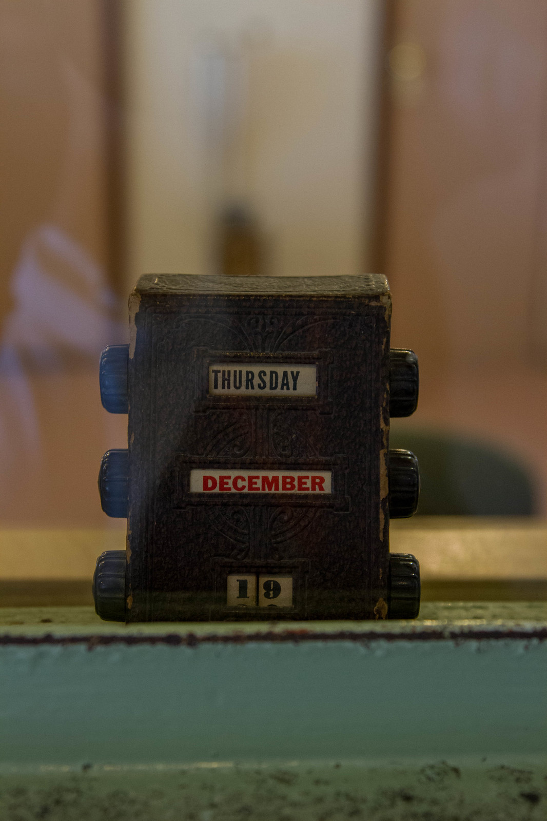 Vintage leather perpetual desk calendar set to Thursday, December 19th, behind glass in a dim room.