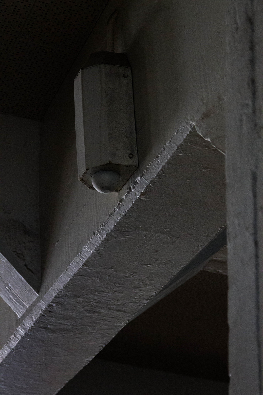 A white octagonal light fixture with a round bulb mounted on a painted concrete wall in a dim stairwell.