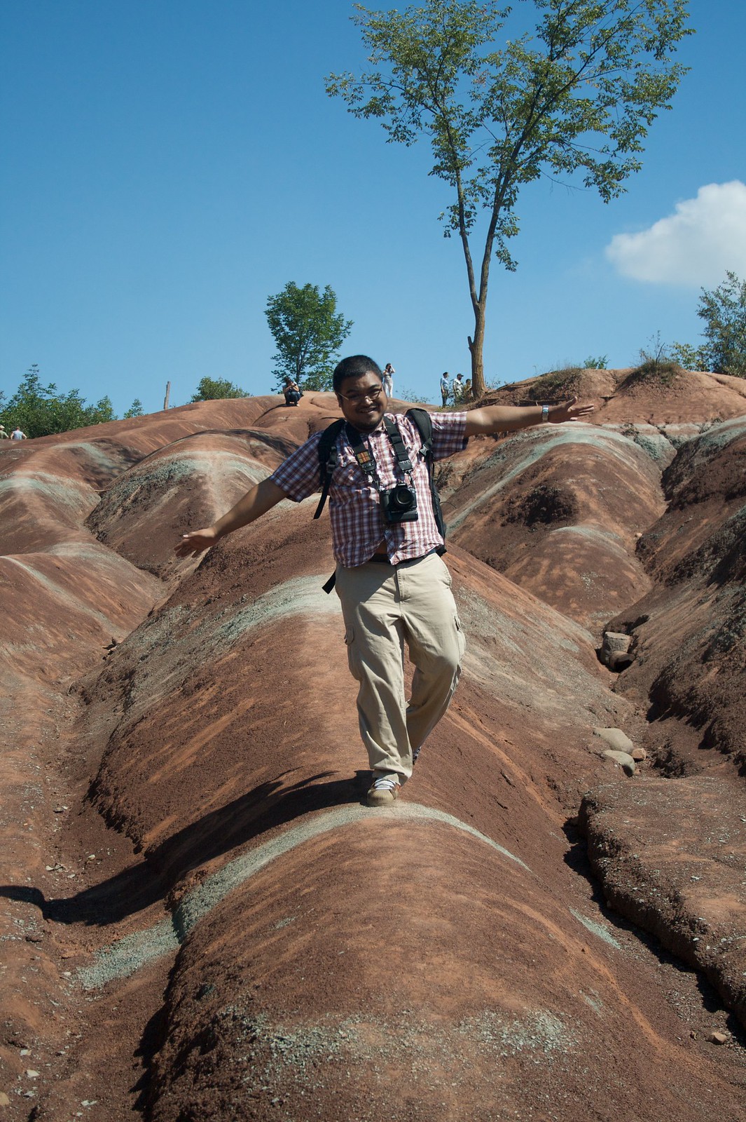 Grinning man with a camera and backpack balances arms outstretched on the red hills of the Cheltenham Badlands.