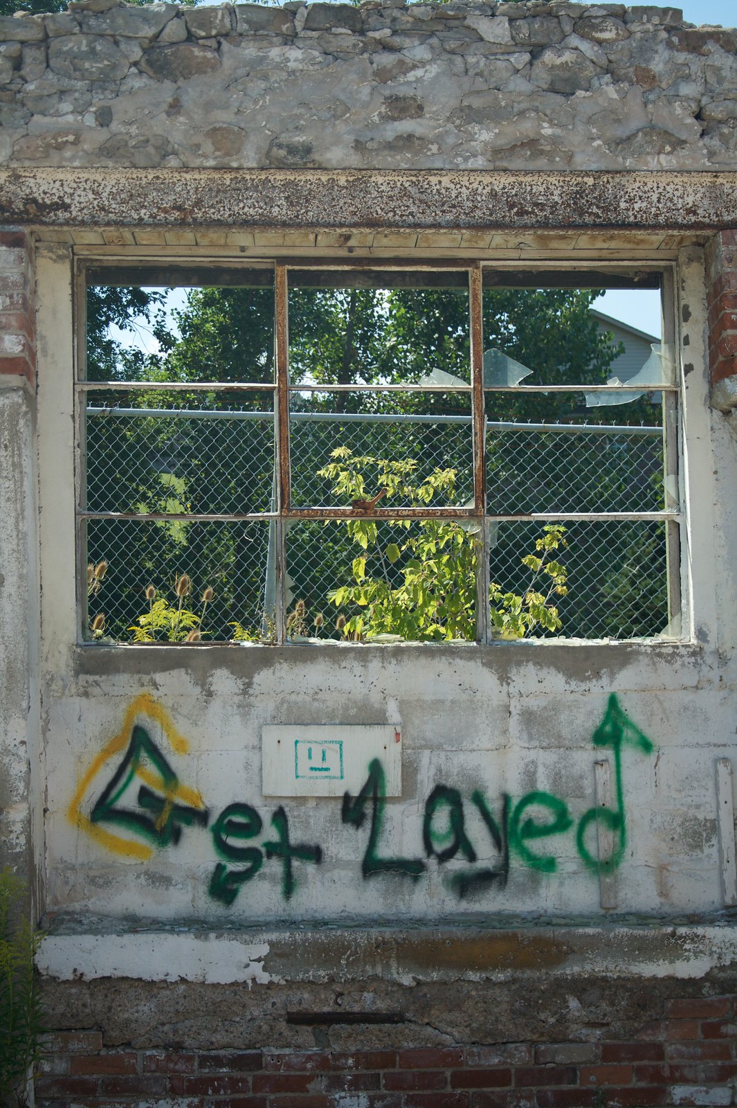 Green graffiti reading "Get Layed" on a crumbling wall below a broken window of an abandoned building.