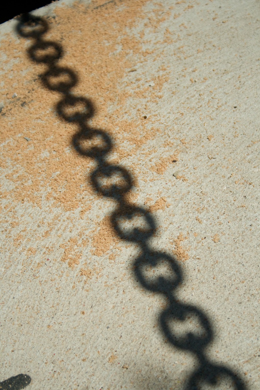 Shadow of a chain's linked rings cast diagonally across a sandy concrete surface.