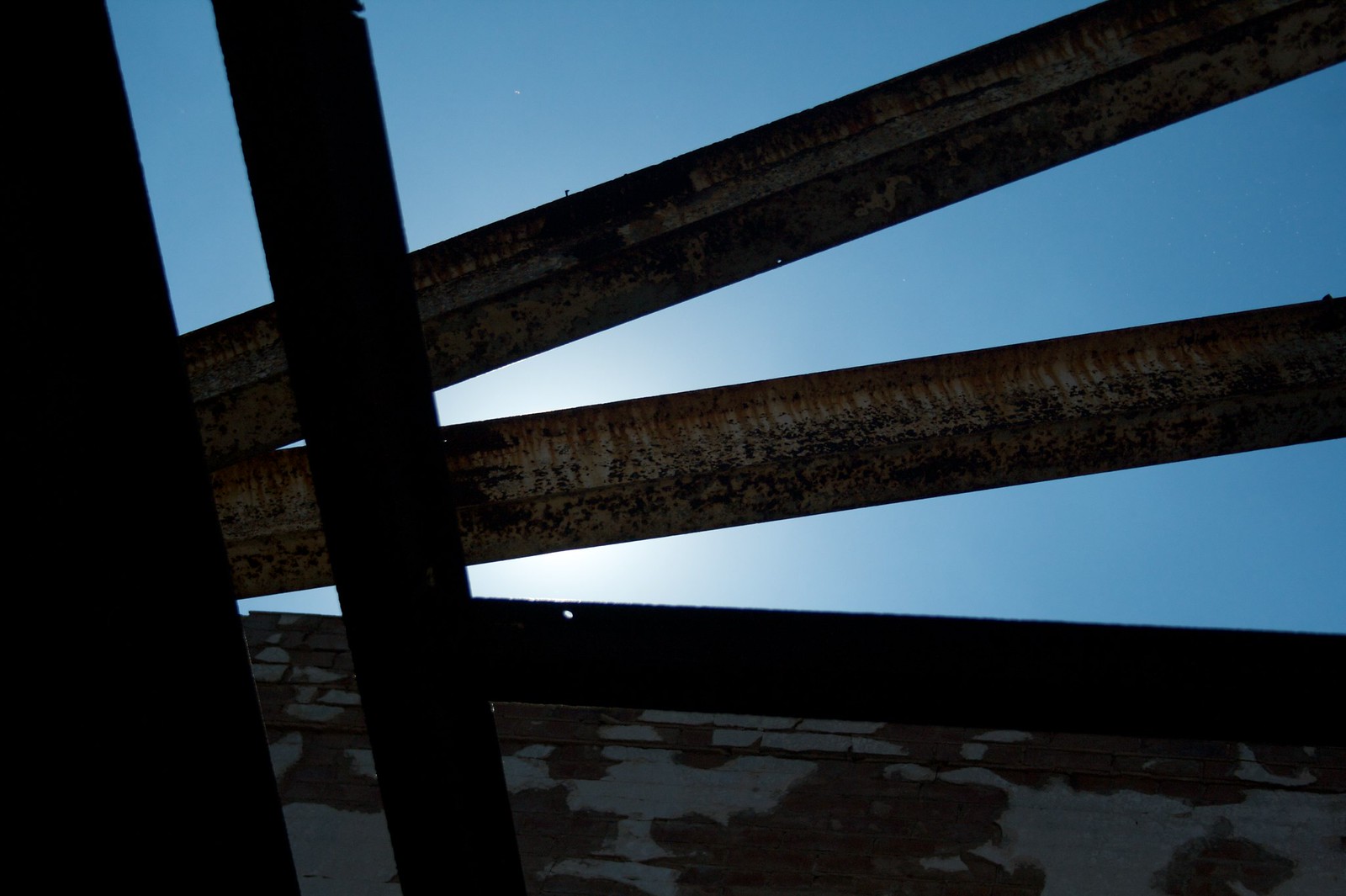 Rusted steel girders of a roofless building silhouetted against a blue sky, sun glowing behind them