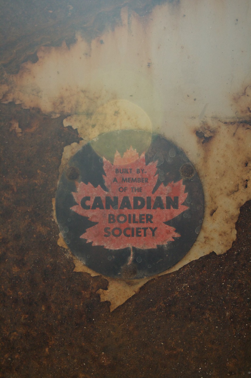 Weathered maple-leaf decal reading "Built by a member of the Canadian Boiler Society" on a rusted metal surface.