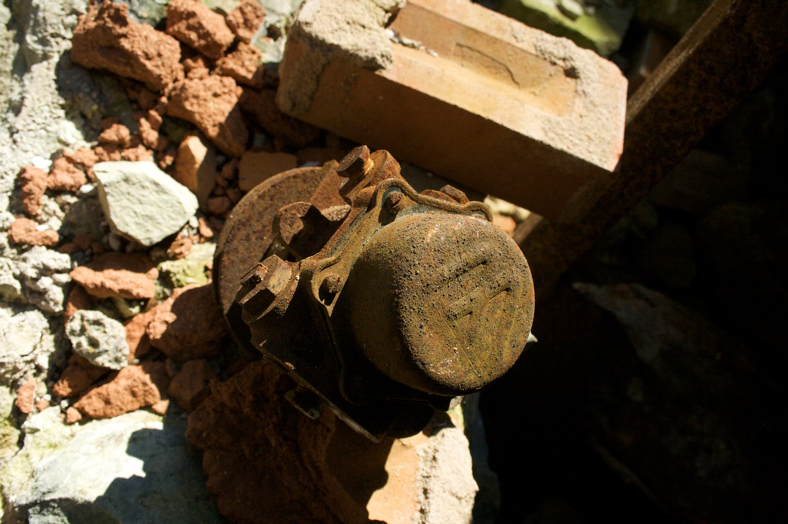 A rusted iron machine part rests among crumbling red bricks and rubble in bright sunlight.