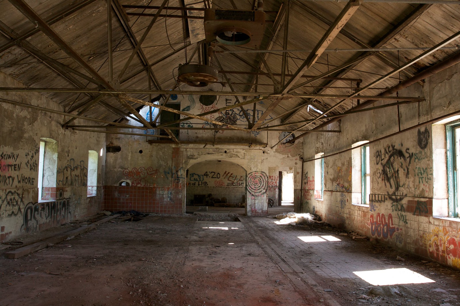 Sunlit interior of an abandoned graffiti-covered building with exposed roof trusses and a tiled dado wall.