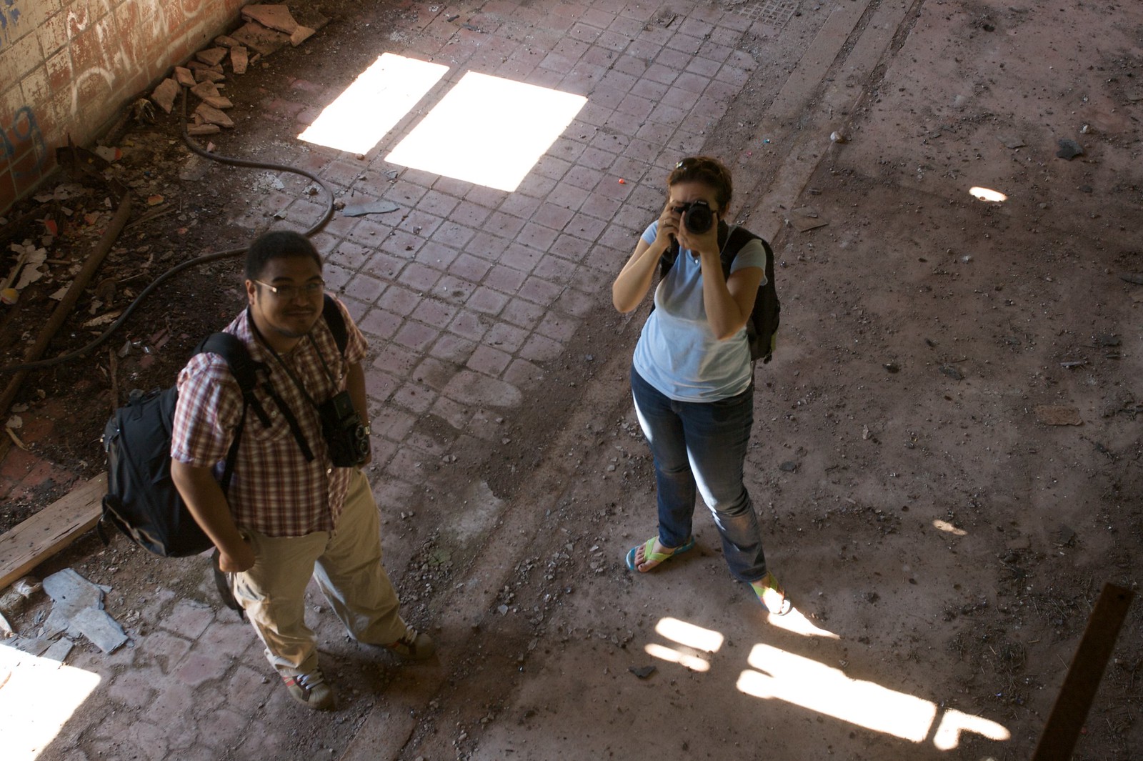 Two photographers with cameras and backpacks stand on the debris-strewn floor of an abandoned building, one aiming her camera upward.
