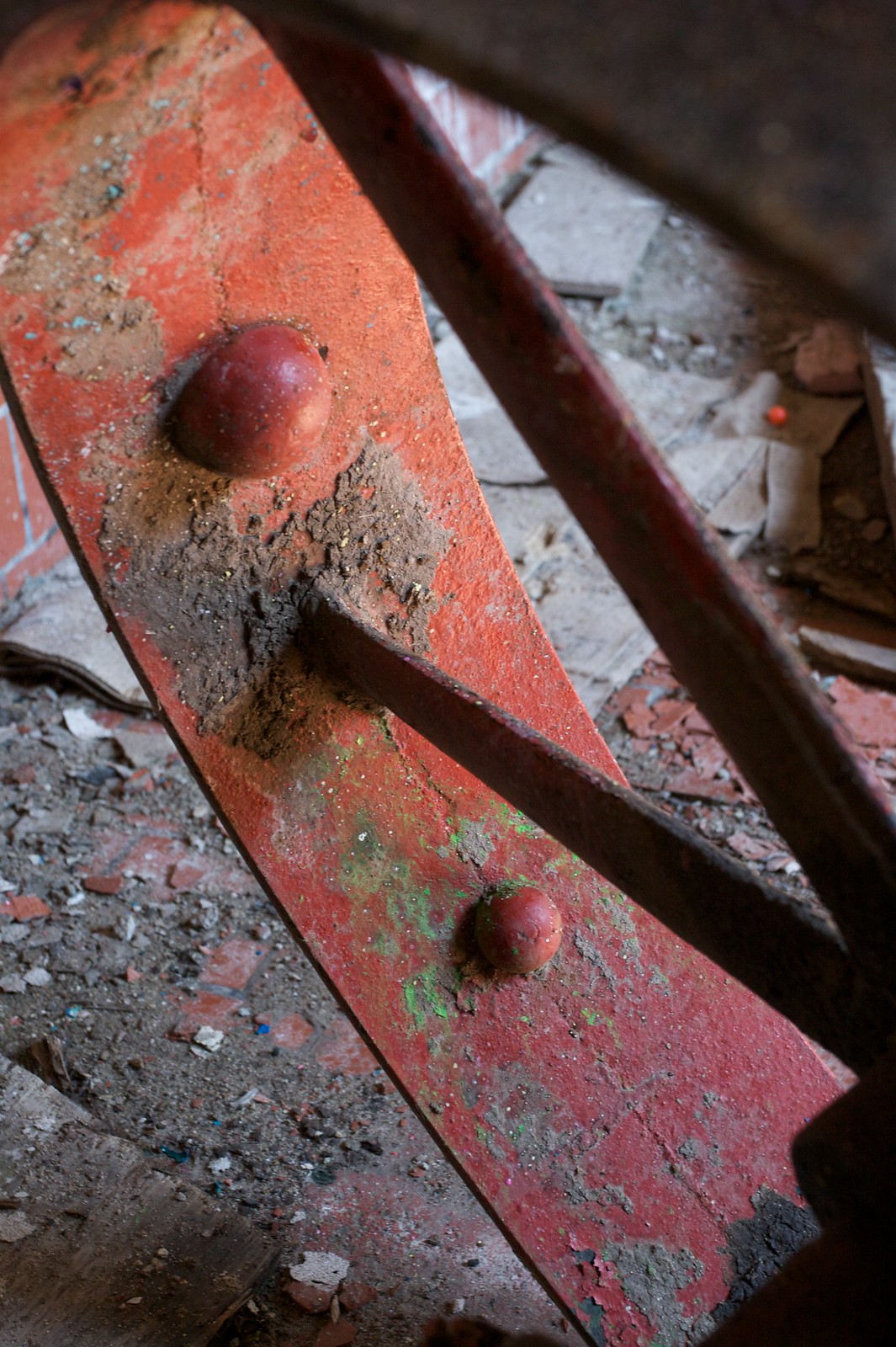 Weathered red barber chair base with two rounded bolt heads, flecked with paint on a debris-strewn floor.