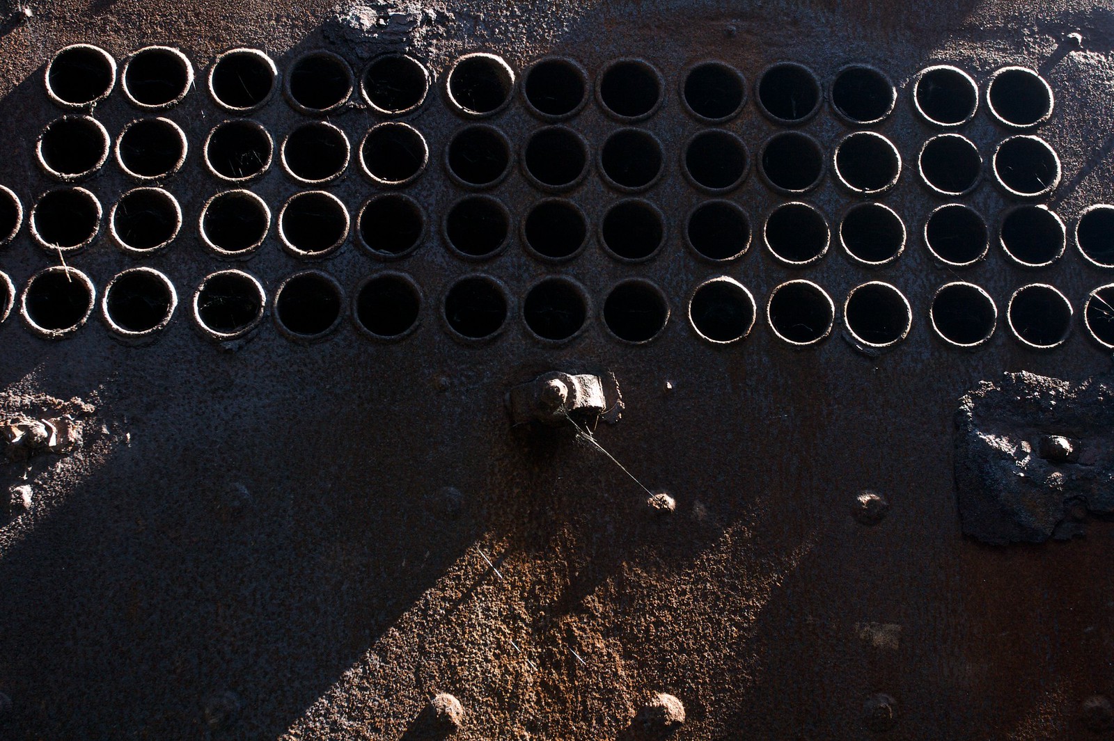 Rows of dark circular boiler tube holes in a rusted steel plate lit by raking light