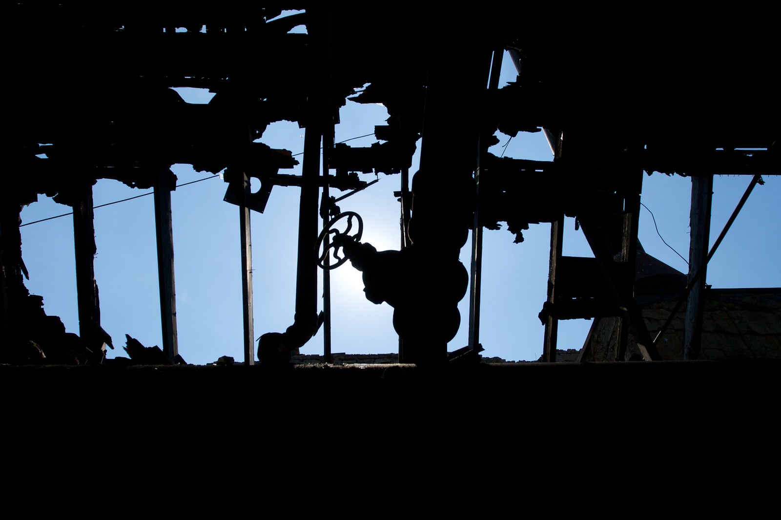 An old valve wheel and industrial pipes silhouetted against blue sky through a derelict factory's decaying roof.