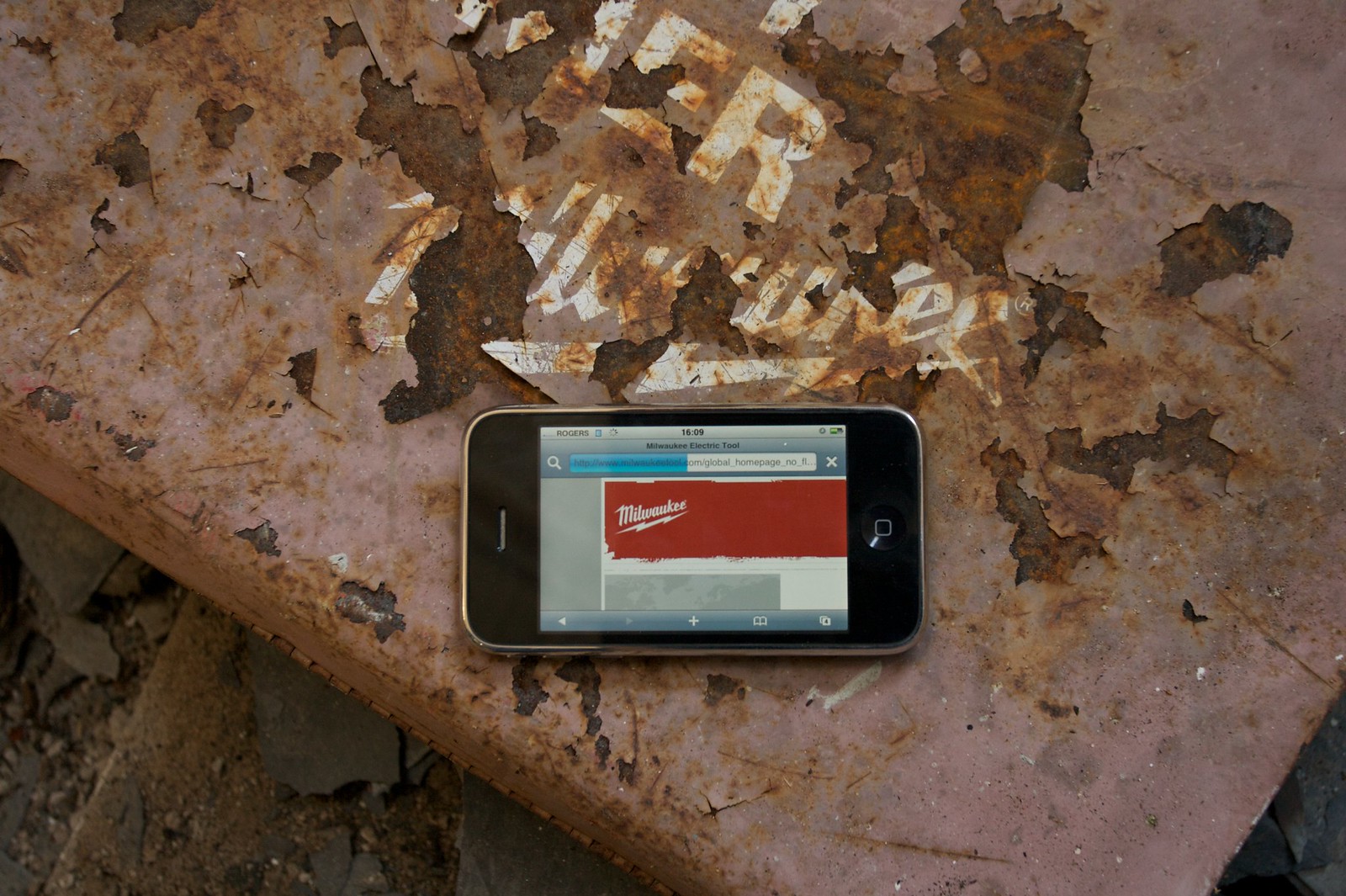 An iPhone showing the Milwaukee tool website rests on a rusted, paint-peeled Milwaukee sign.