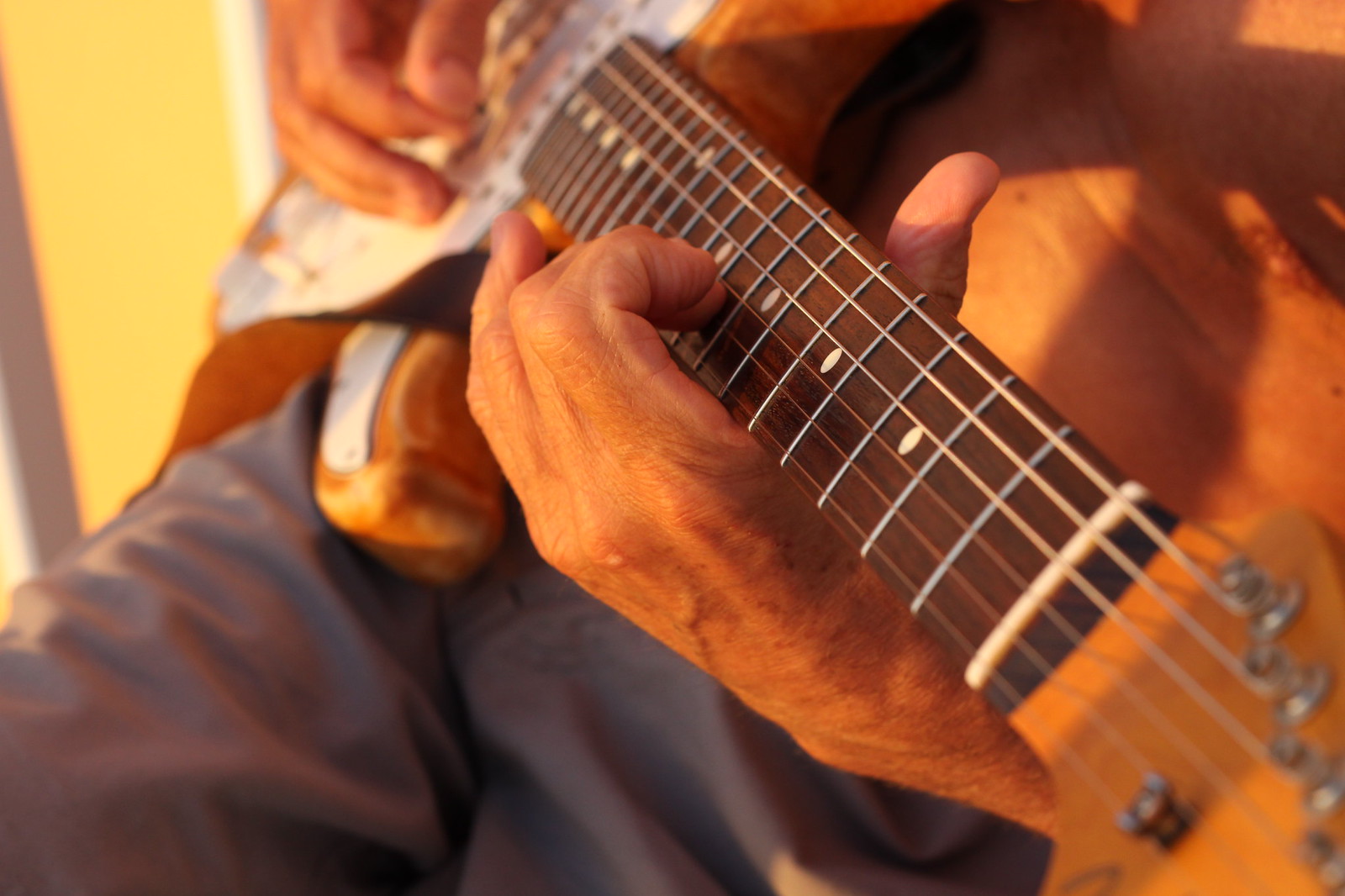 Weathered hands play an electric guitar, fingers pressing frets in warm golden sunset light.