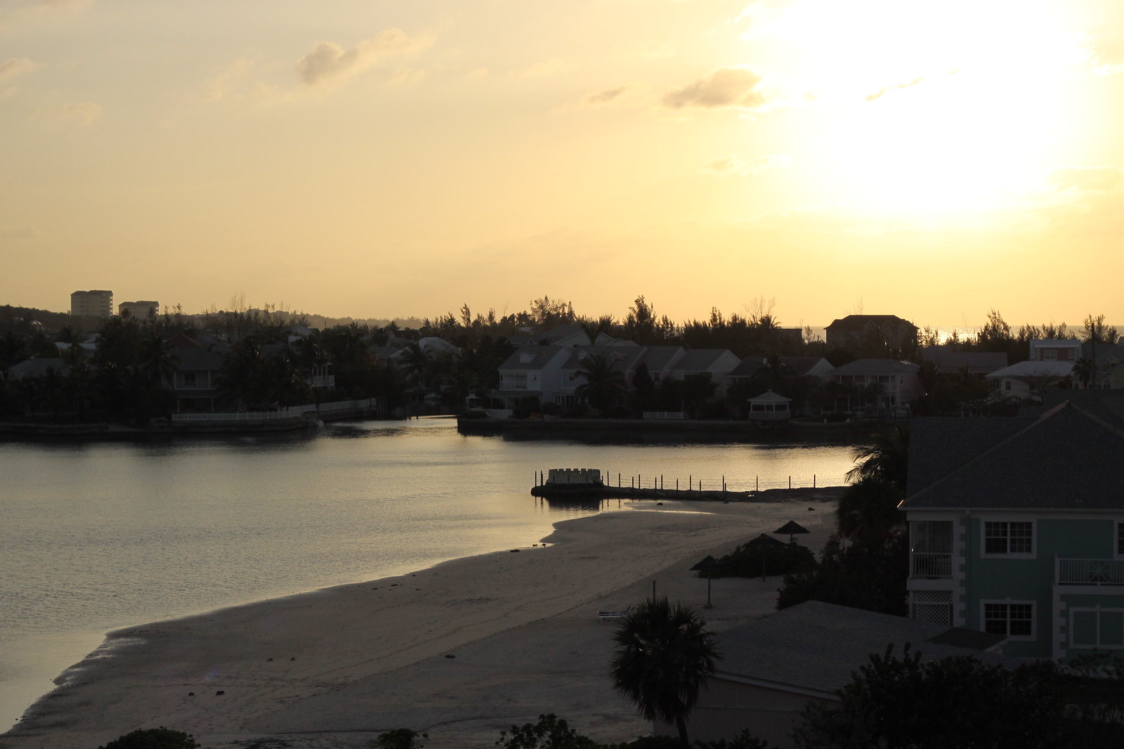 Golden sunset over a calm bay with a sandy beach, dock, and waterfront homes fringed by palm trees.