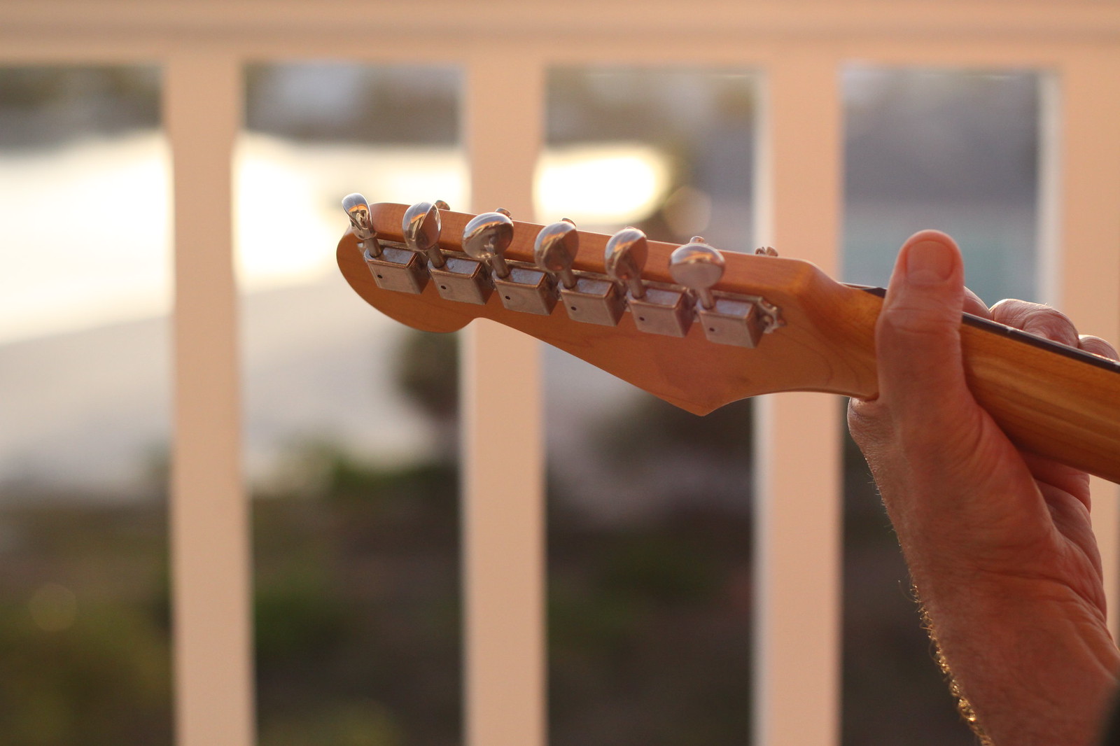 Backlit electric guitar headstock held up against a sunset, tuning pegs glowing, blurred railing behind.