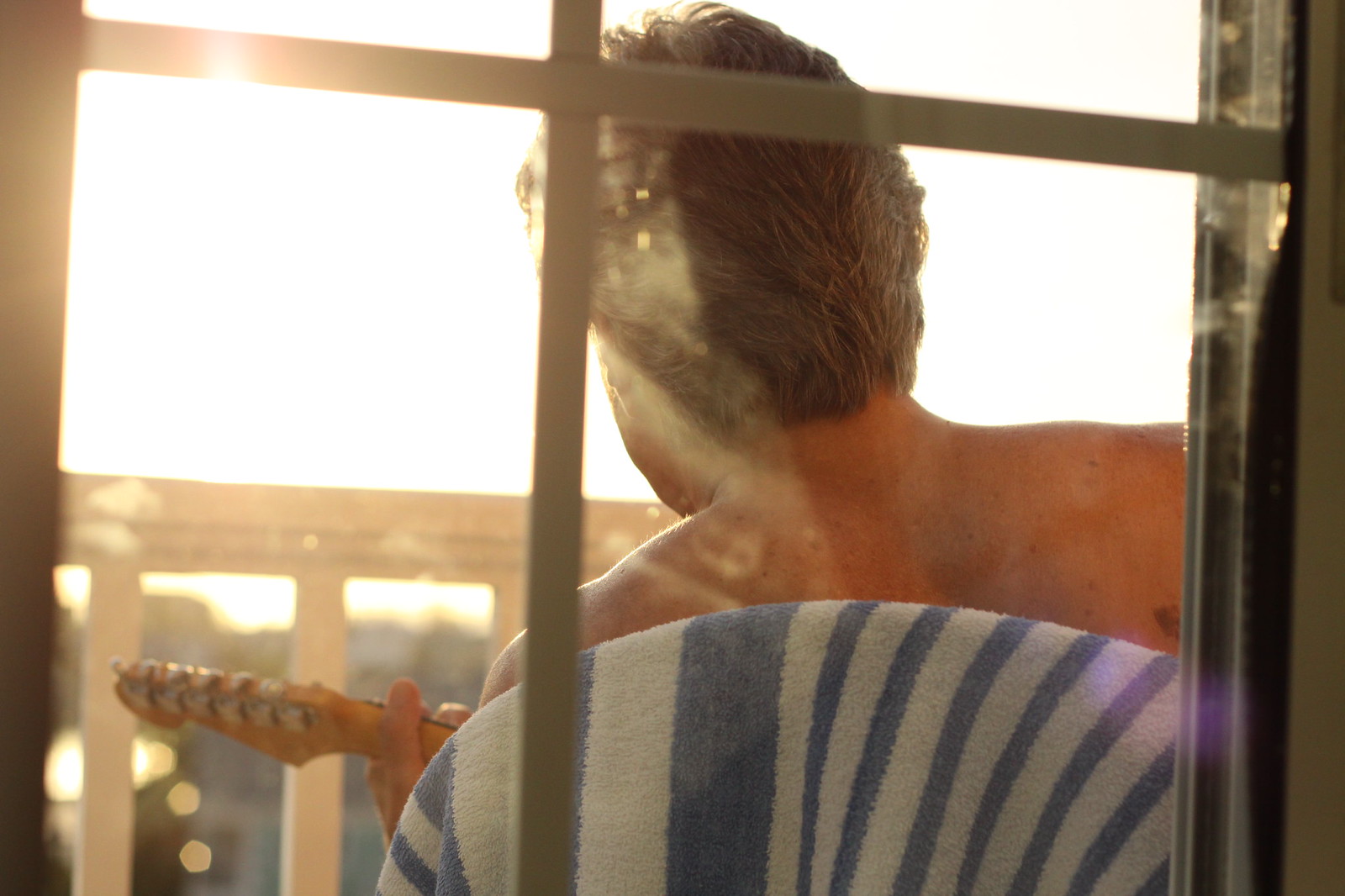 Shirtless man with a striped towel over his shoulder holds an electric guitar, facing a sun-drenched window at golden hour.