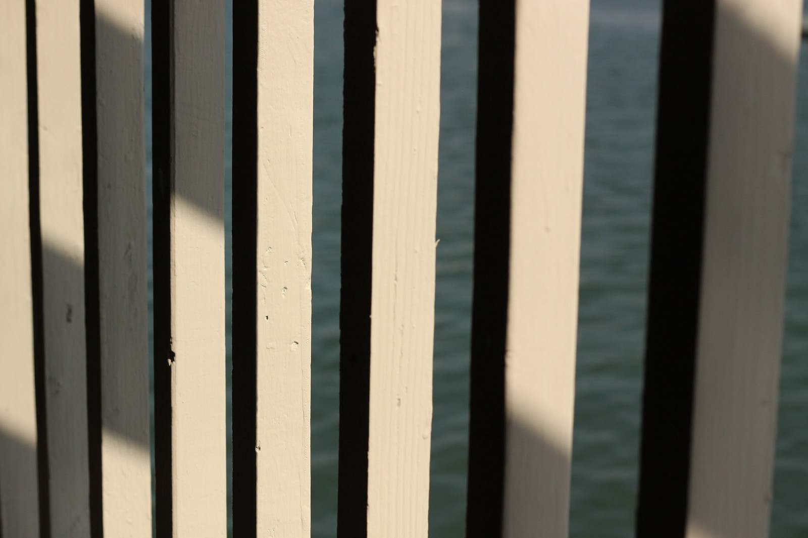 Sunlight and shadow falling across a white painted wooden railing overlooking calm blue-green water.