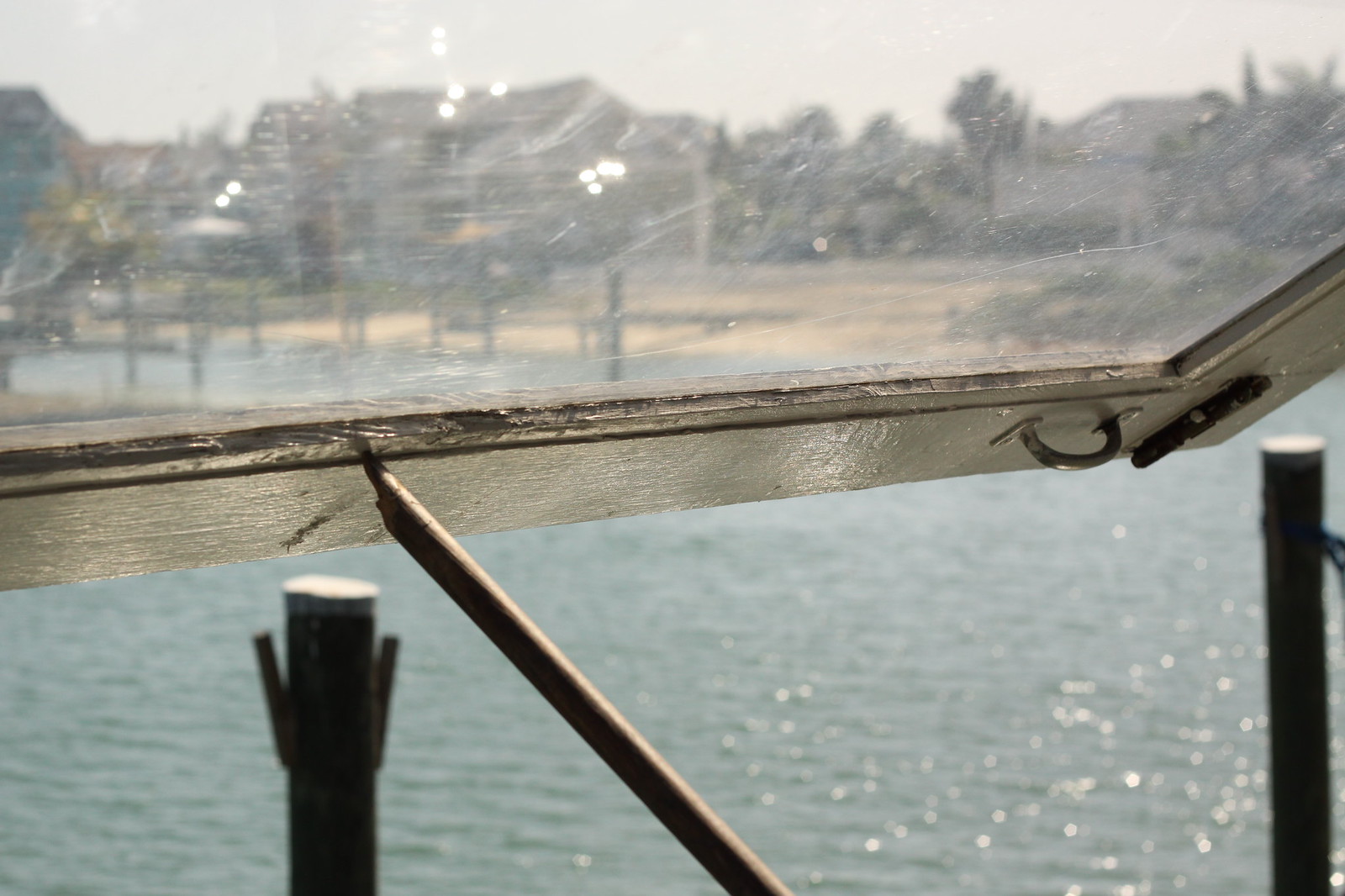 View through a scratched, sun-flecked boat window of weathered wooden dock pilings and calm turquoise water, with a hazy shoreline beyond.
