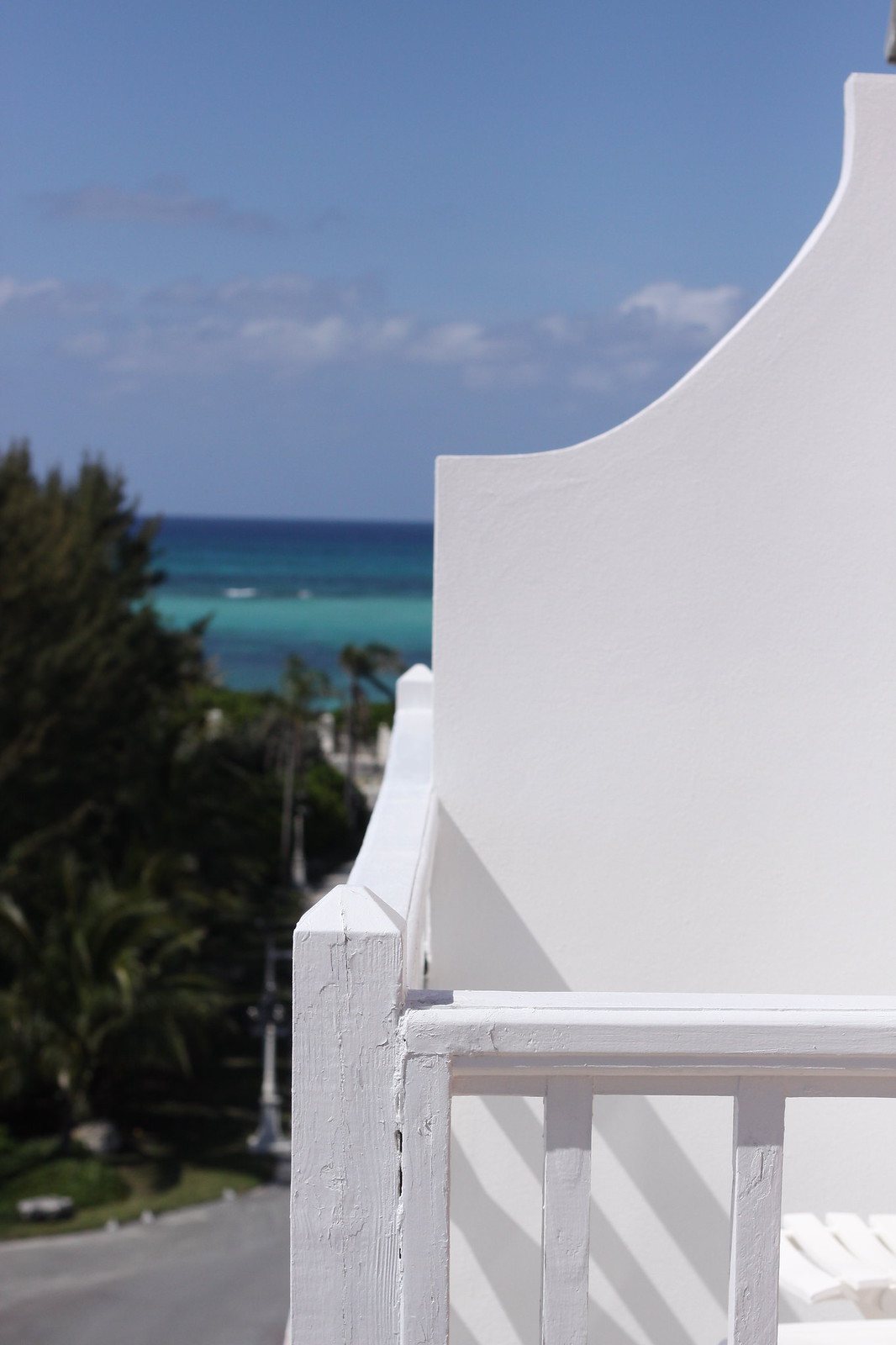 Curved white Bermuda gable and balcony railing overlooking palm trees and turquoise ocean under a blue sky.