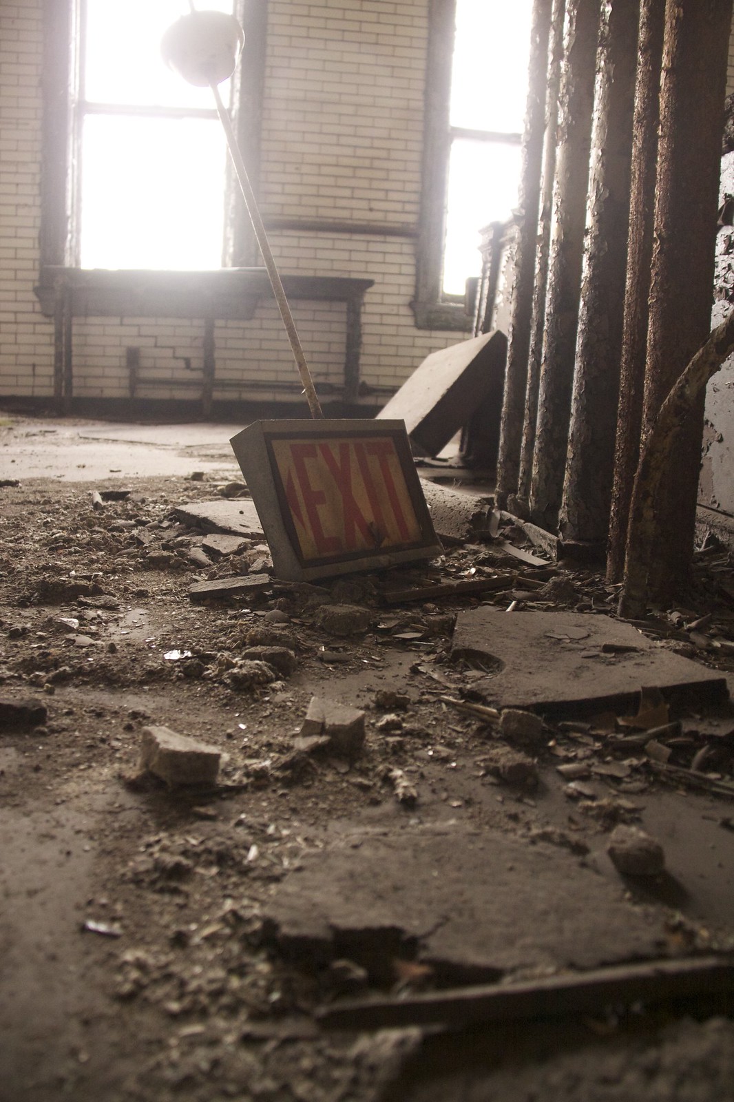 A toppled illuminated "EXIT" sign lying amid rubble and peeling paint in an abandoned tile-walled room.