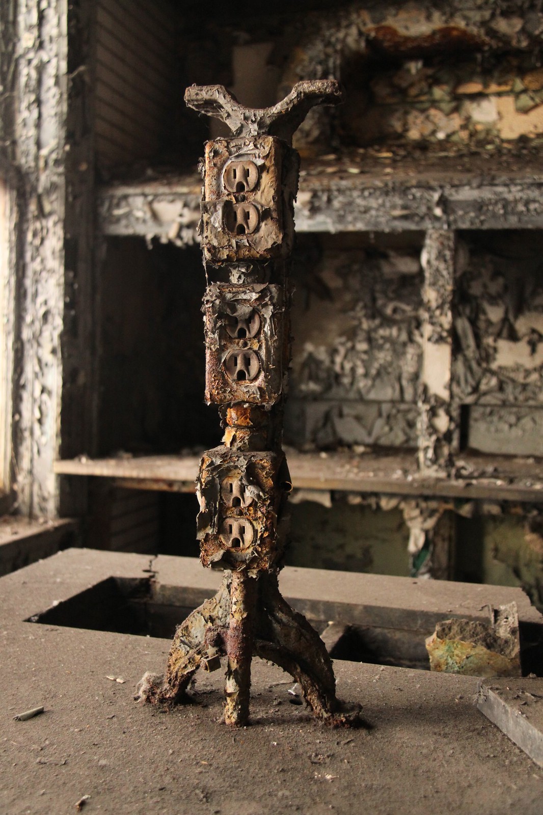 A rusted, fire-scorched tower of stacked electrical outlets stands in a burned-out, decaying industrial office.