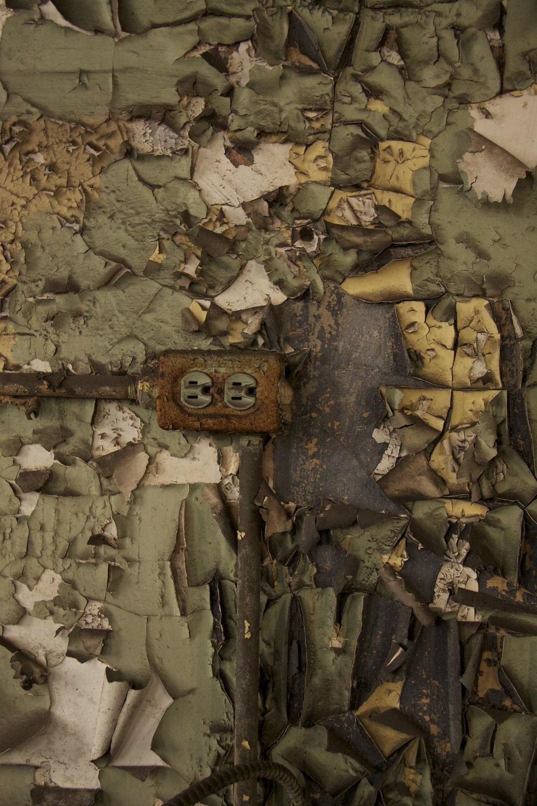 Rusted electrical outlet on a wall of thickly peeling green and yellow paint in an abandoned building.