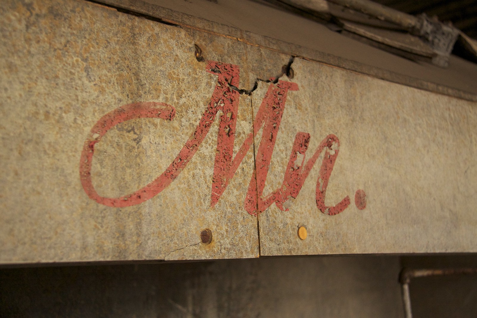 Faded red hand-painted cursive "Mr." lettering on a weathered, peeling sign in an abandoned industrial building.