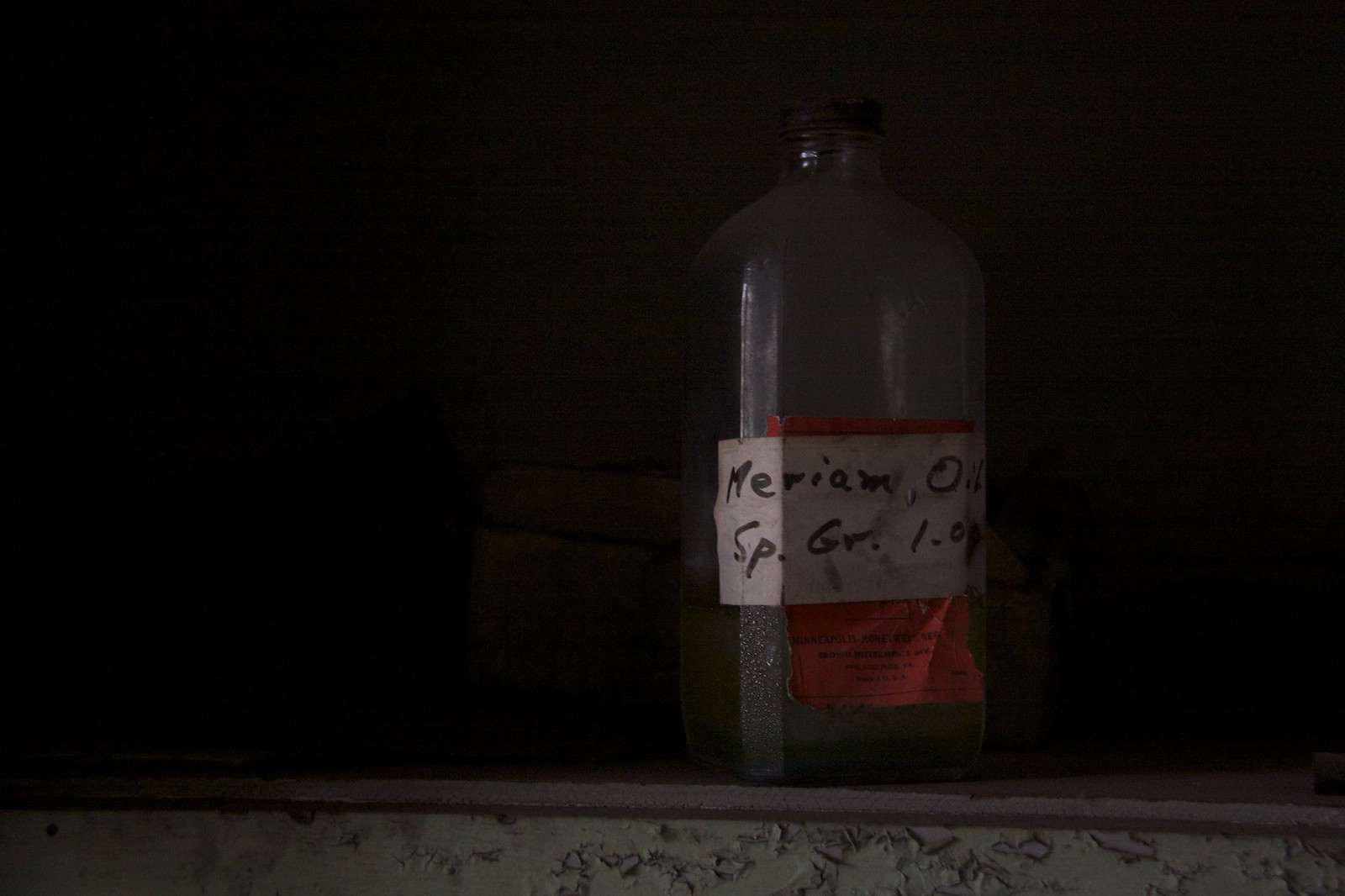 Old glass bottle on a shelf in a dark room, handwritten label reading "Meriam Oil, Sp. Gr. 1.0
