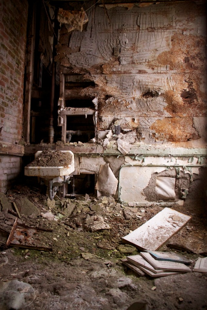 A rusted porcelain sink hangs on a decaying wall in an abandoned office strewn with rubble and scattered papers.