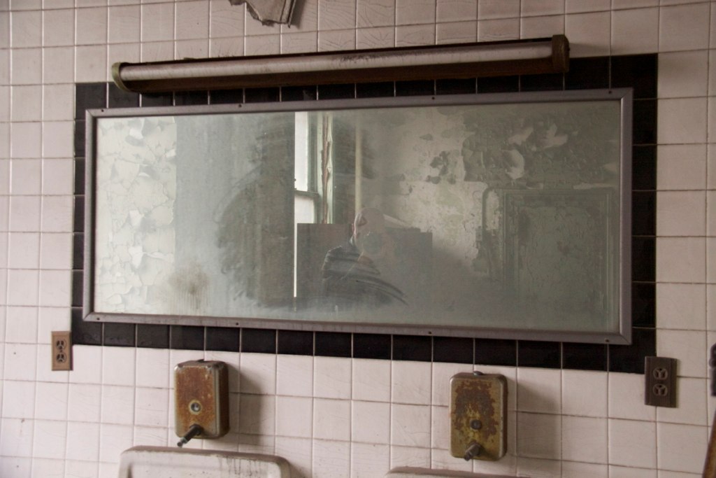 A ghostly figure reflected in a grimy mirror above rusted soap dispensers in an abandoned tiled restroom.