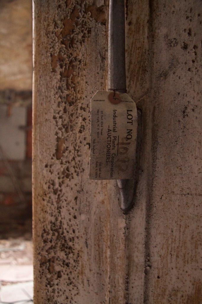 Weathered auction tag reading "LOT NO." and "Industrial Plants Corporation Auctioneers" wired to a rusted metal lever.