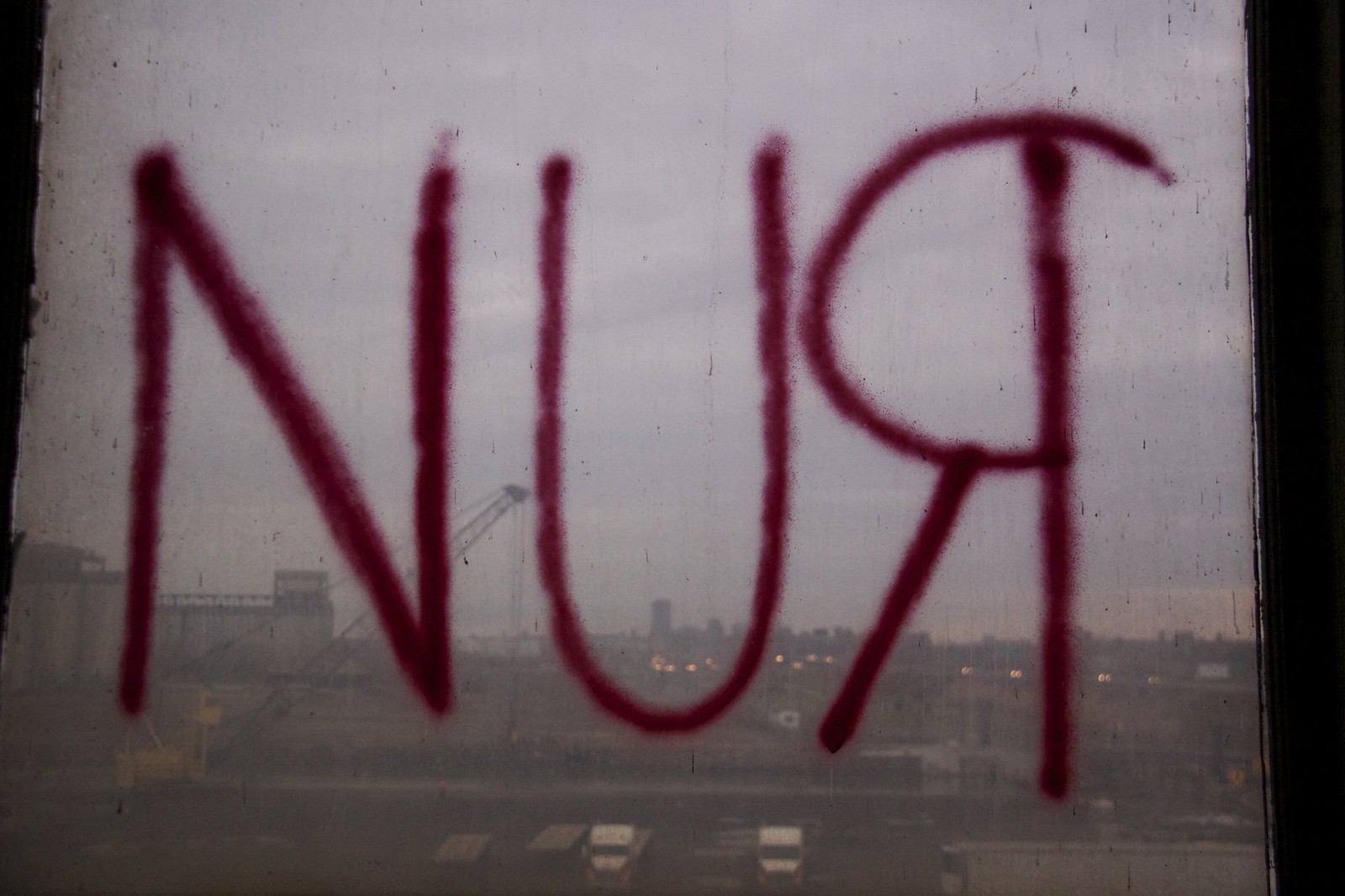 RUN" spray-painted in red on a grimy window overlooking a hazy industrial skyline at dusk.