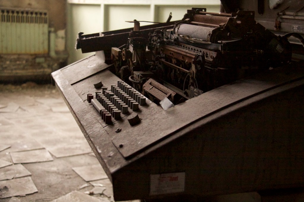 Rusted antique adding machine or typewriter sitting on a wooden desk in an abandoned, debris-strewn room