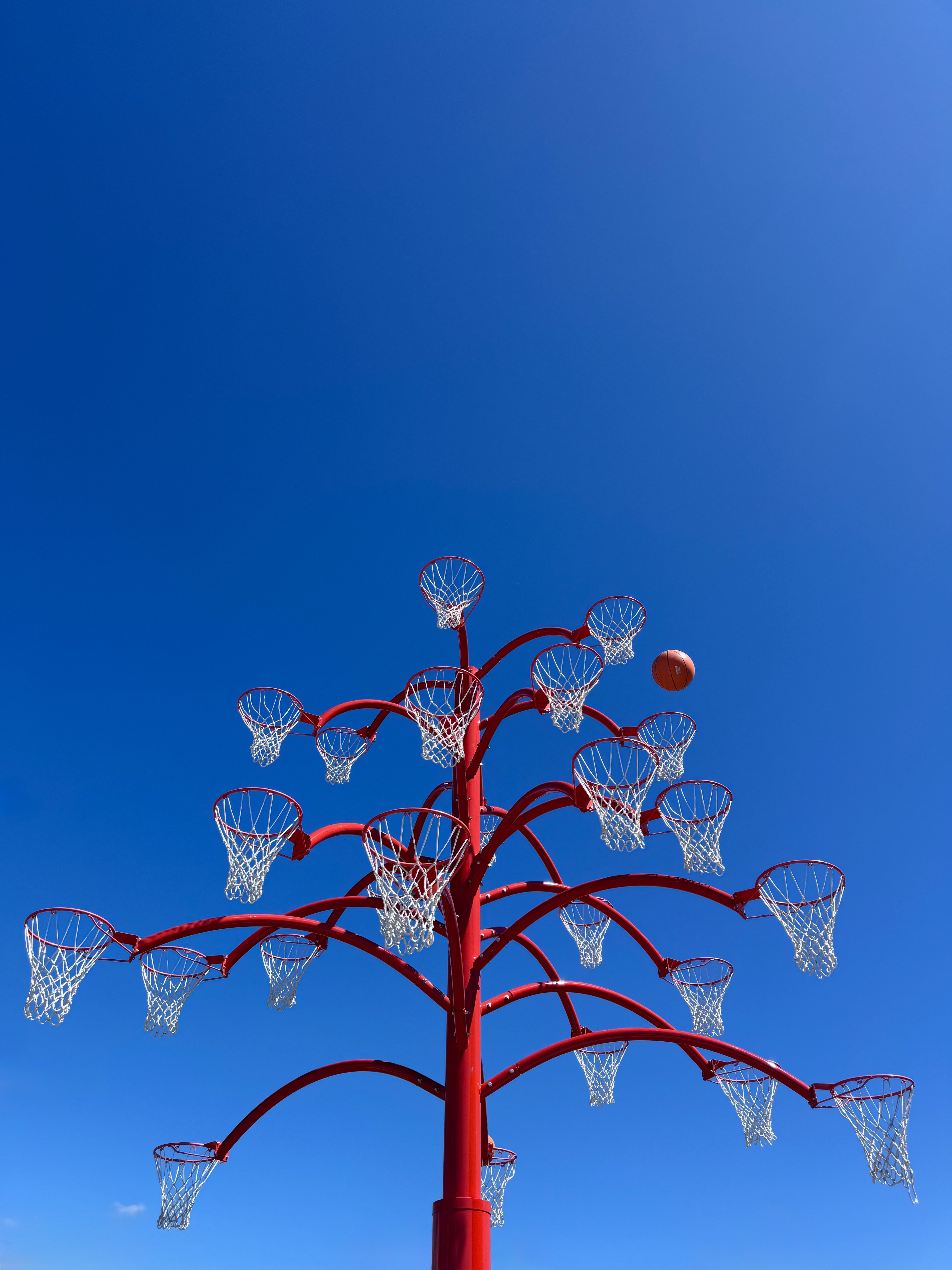 Red tree-like sculpture of many basketball hoops with a floating basketball against a clear blue sky