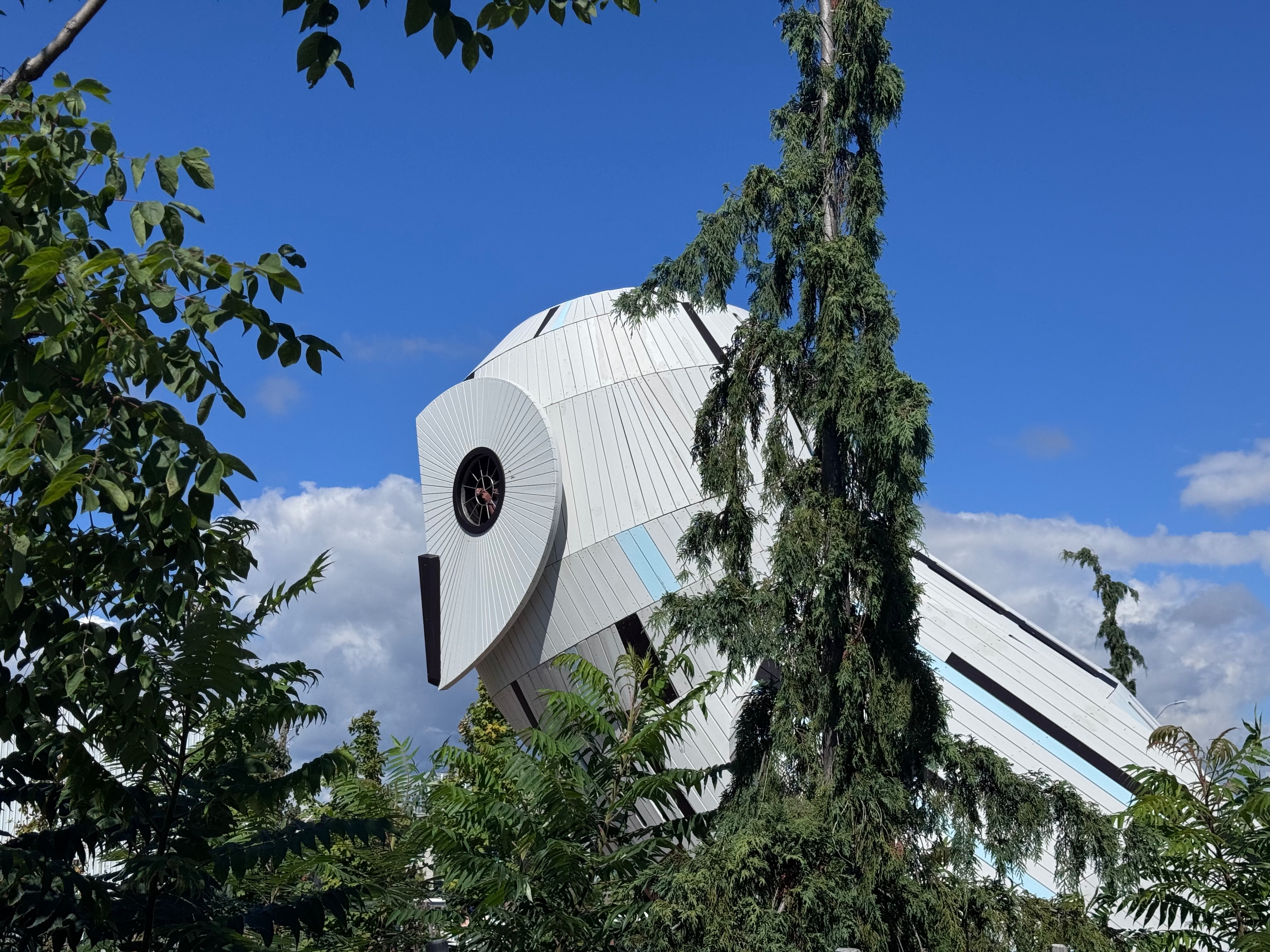 A giant wooden owl-shaped sculptural building with a large round windows for eyes rises behind lush green trees under a bright blue sky.